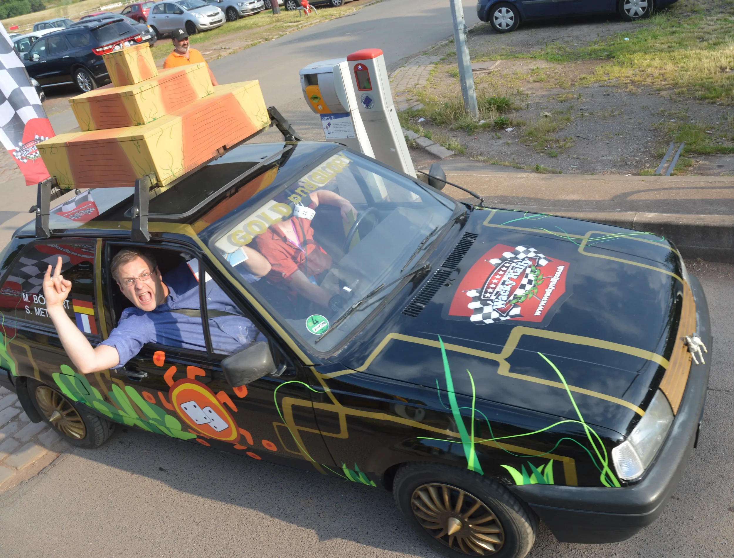 A decorated racing car with a driver and passenger inside. The car has colorful graphics, a large checkered flag logo on the hood, and a rack of large, colorful boxes on the roof. The driver appears excited, making a victory sign with his hand. The v