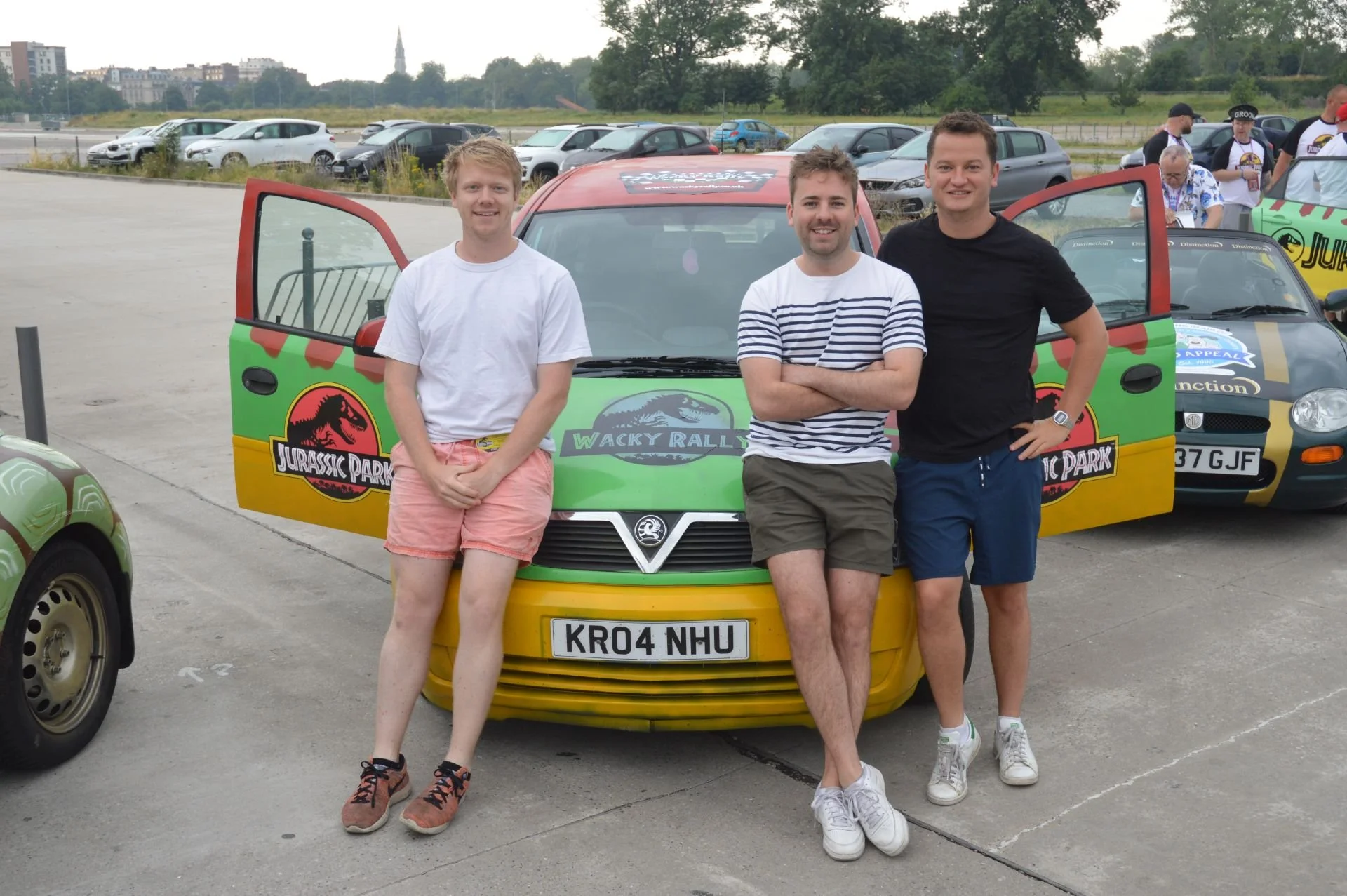 Three young men stand in front of a car with a Jurassic Park and Jurassic World theme at an outdoor parking lot, with other cars and people in the background.