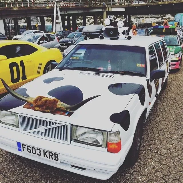 A parking lot with a white station wagon decorated to resemble a cow, featuring black spots, cow horns, and a cow tail, with other cars and a person in the background.