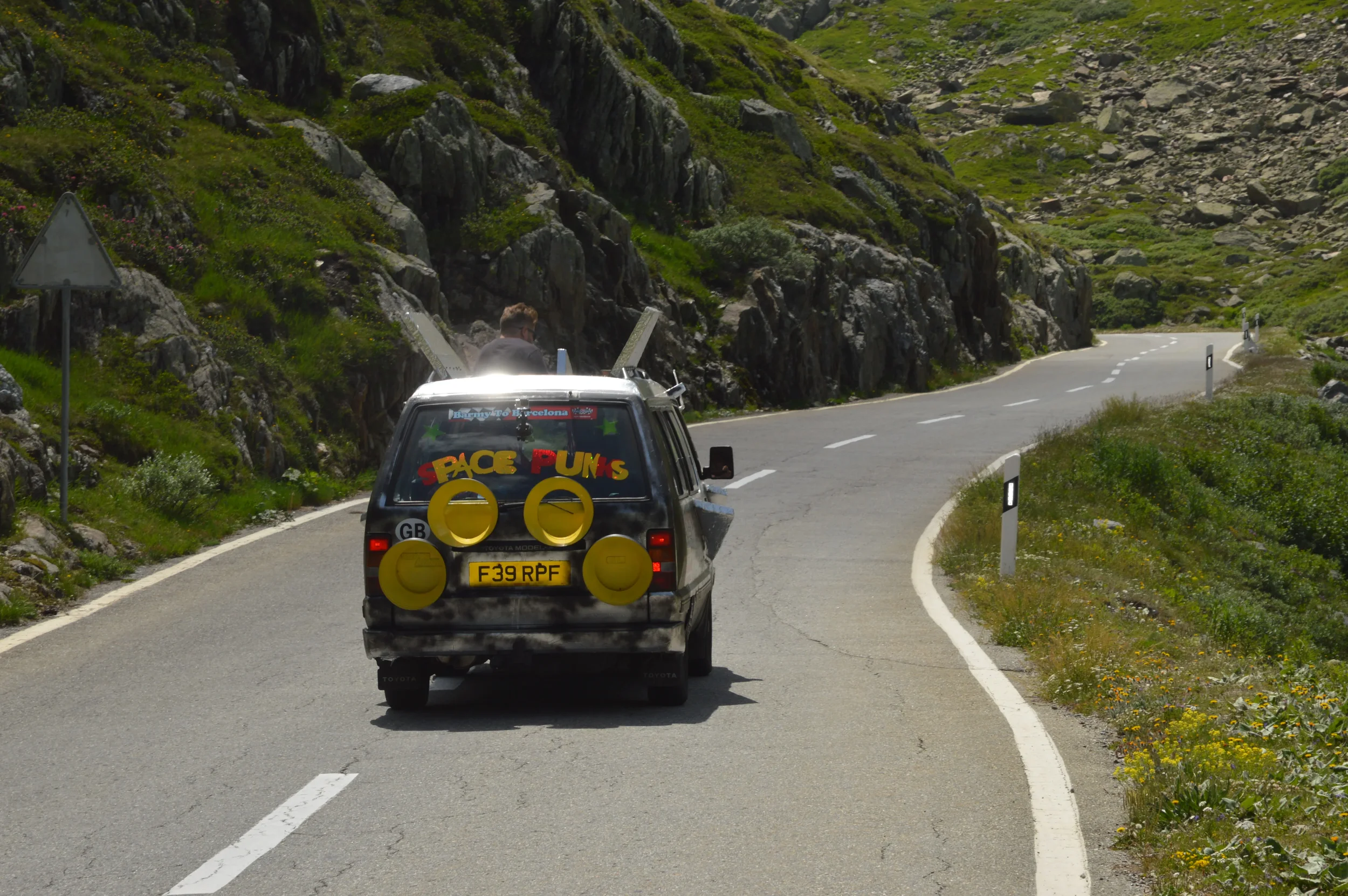 A black SUV with colorful stickers and large yellow speakers on the back driving on a winding mountain road with rocky terrain and green vegetation on each side.