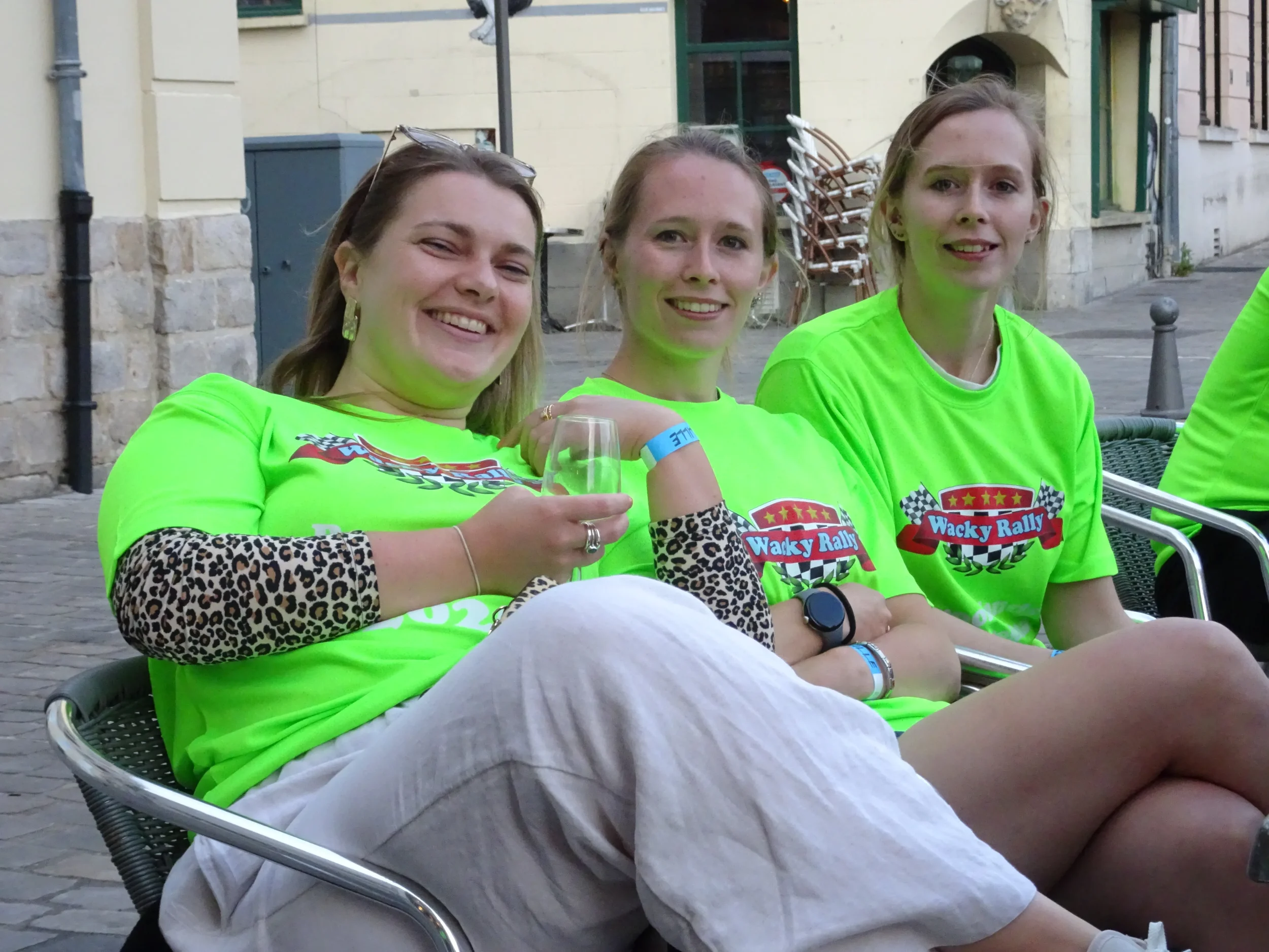 Three women sitting outdoors on metal chairs, smiling, wearing bright green T-shirts with 'Wacky Rally' logo, one holding a glass of drink.