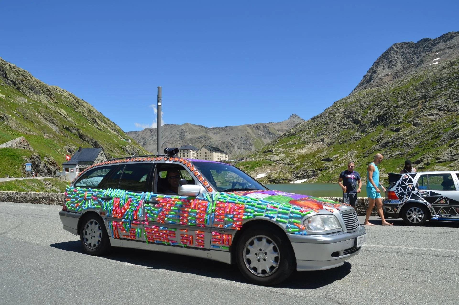 A silver station wagon decorated with colorful plastic noodles and bottles, parked on a mountain road under a blue sky. Three people are nearby, one looking at the car, others walking, with mountainous scenery and a lake in the background.