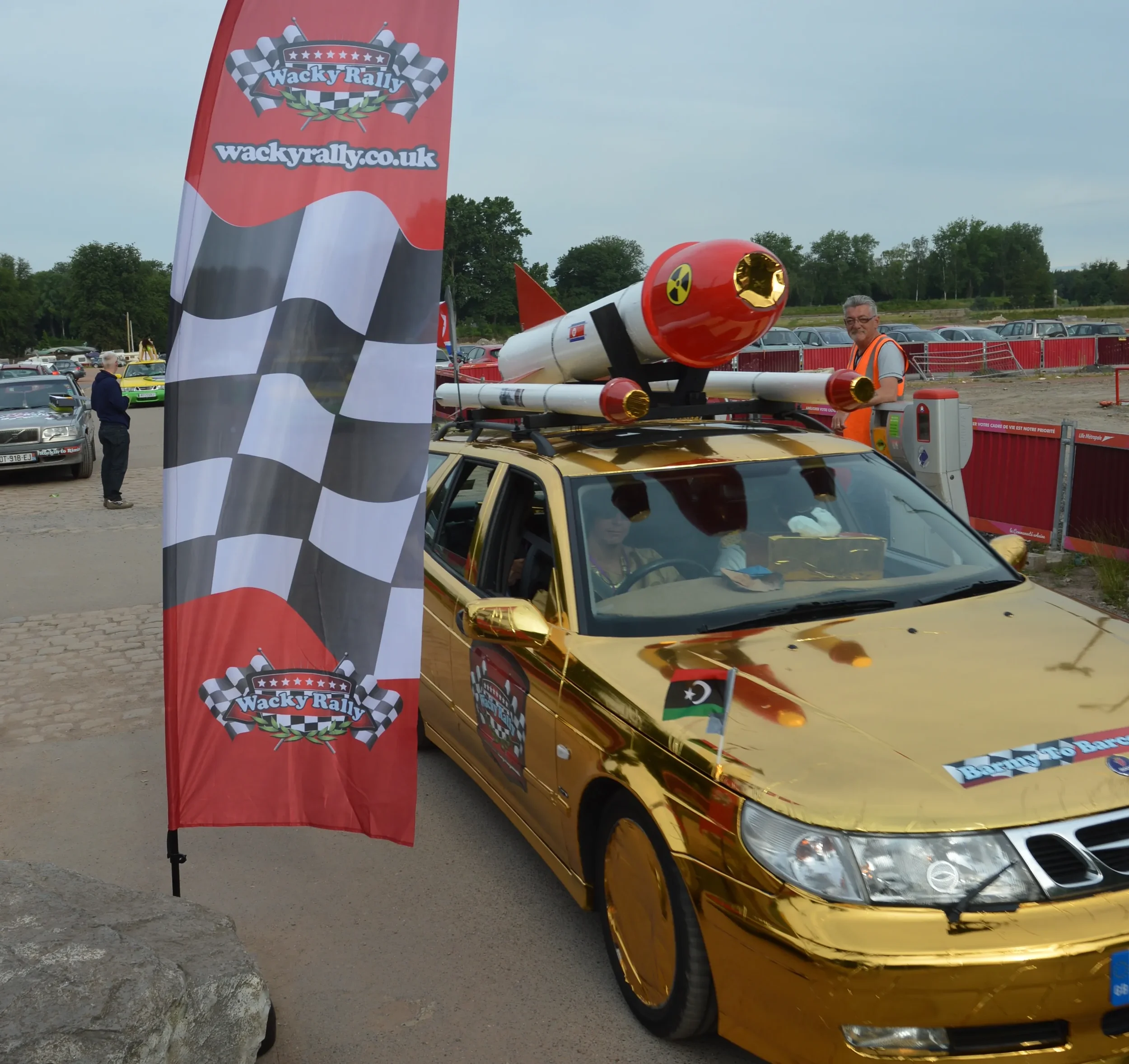 A gold-colored car decorated with a Libya flag and rally stickers, parked at a rally event. The car has a rocket model on its roof and is near a red Wacky Rally banner. People are in the background, some taking photos and others observing.
