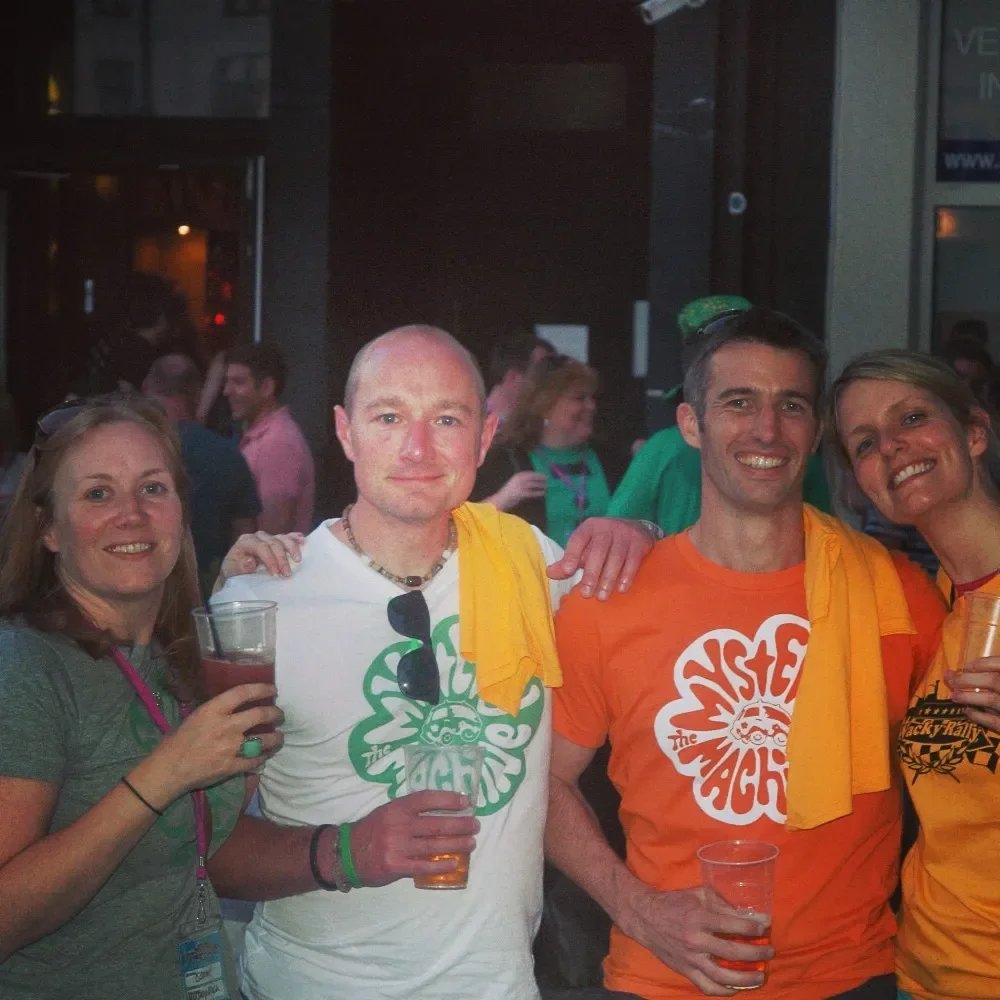 Four friends gathered at a party, smiling and holding drinks, with others dancing in the background.