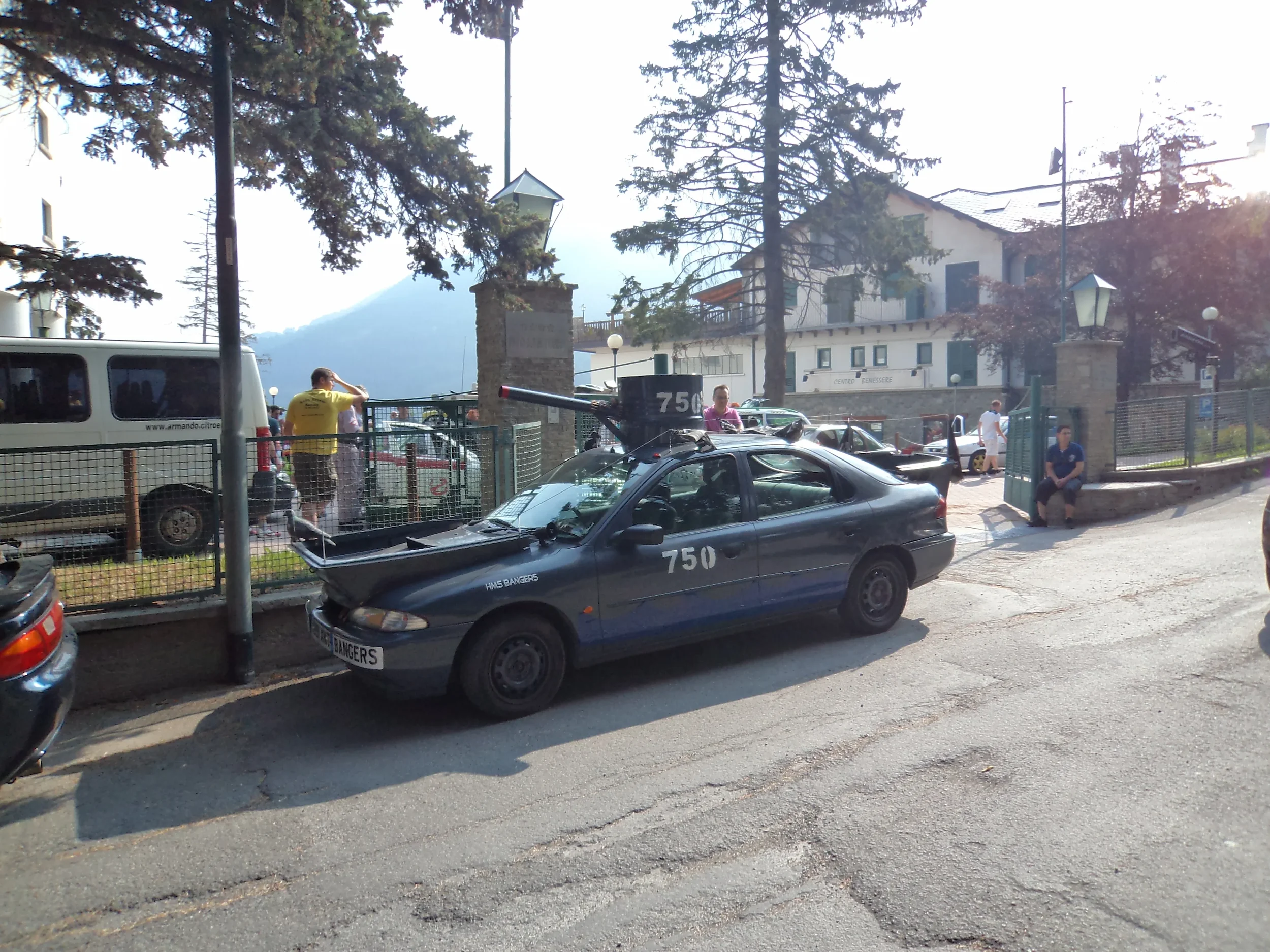 A dark gray car modified for military use with a mounted gun on the roof parked on a street. Several people are nearby, some in front of a fenced area with trees and buildings in the background, and the sun is shining brightly.