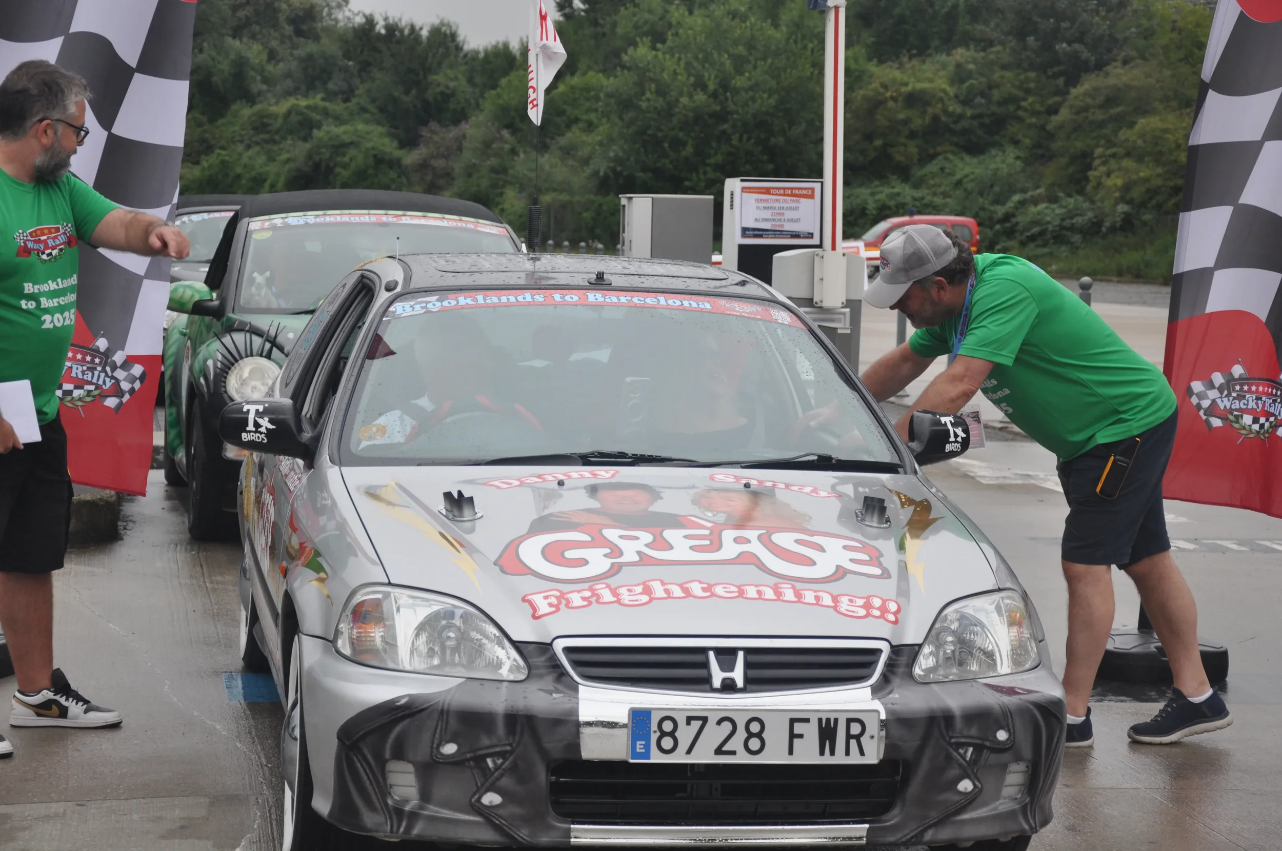 A silver Honda race car with promotional graphics on the hood, two people in green shirts attend to it at a race track, with a second race car behind it. The scene is set outdoors with trees in the background and race banners on either side.