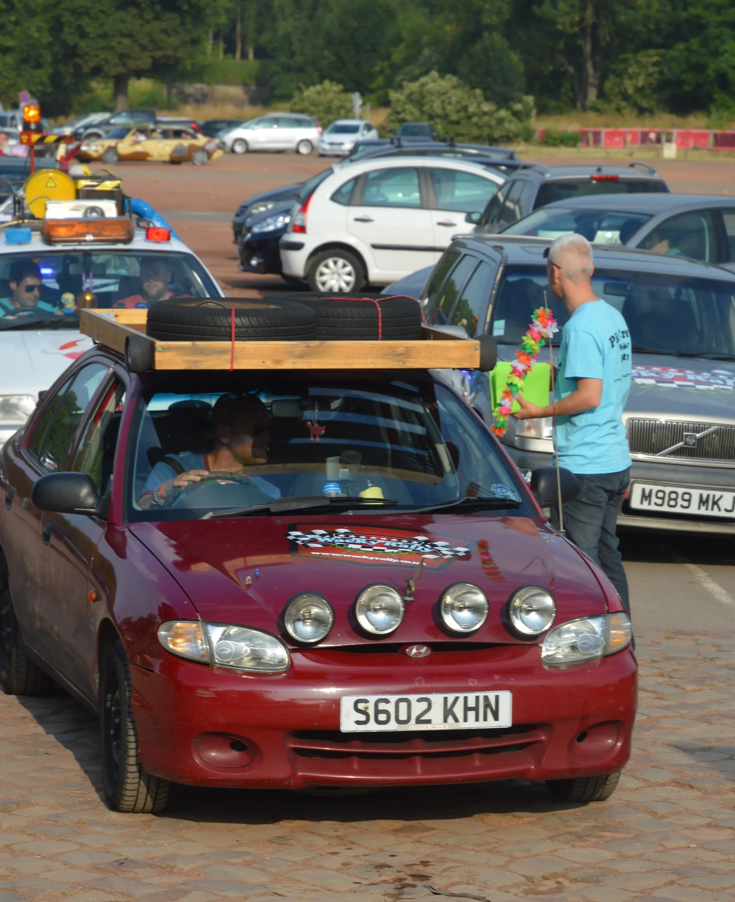 Red car with four large rally-style spotlights on the hood, driving license plate S602 KHN, parked in a busy parking lot with other cars and people, one person standing by the car holding a colorful lei and wearing a blue t-shirt.