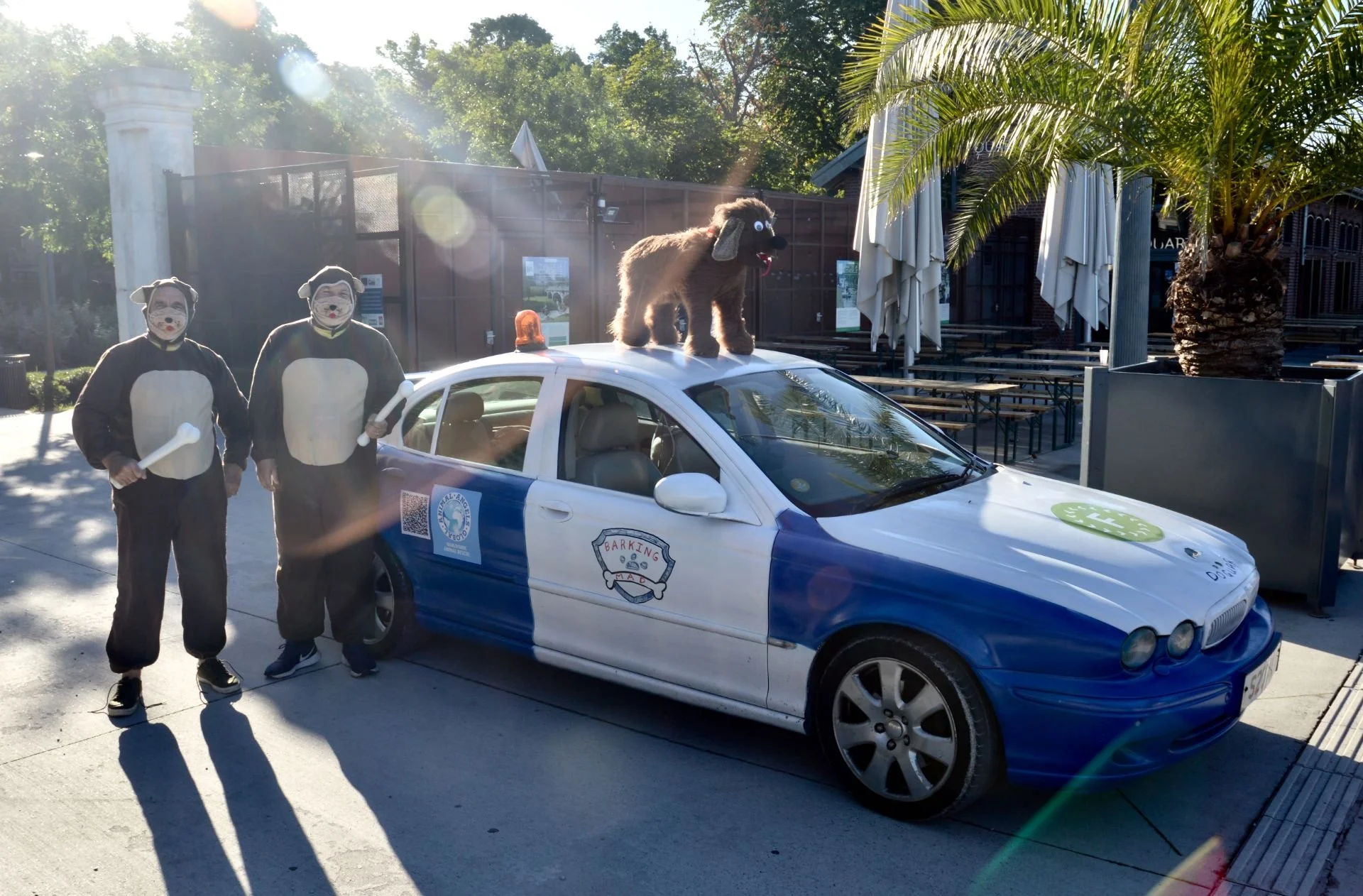 Two people dressed as dogs standing next to a police car with a large dog figure on the roof at an outdoor location with palm trees and outfitted with police and dog-themed decorations.