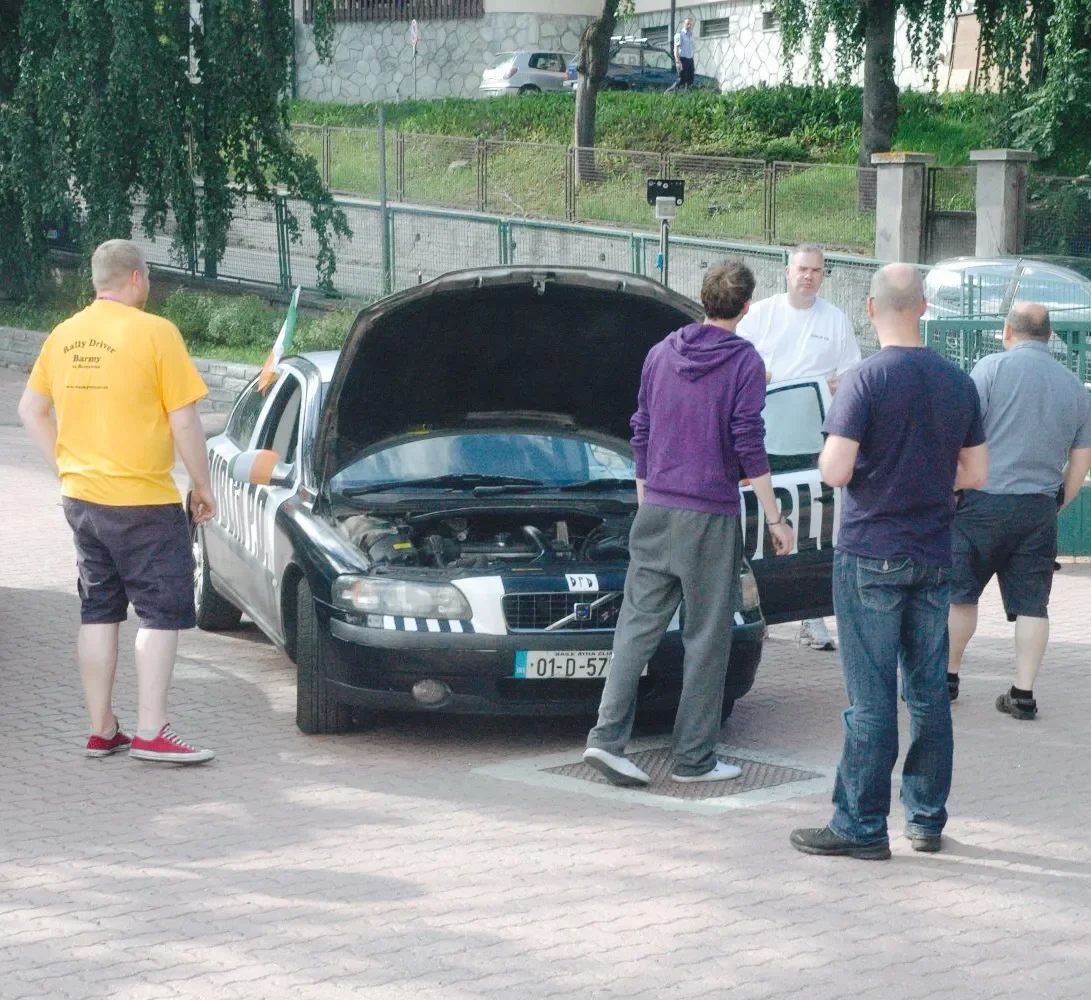 Group of six men gathered around an open hood police car on a paved street, with one man in a yellow shirt, others in casual clothing, and a clipboard, in an outdoor area with some greenery and a stone wall in the background.