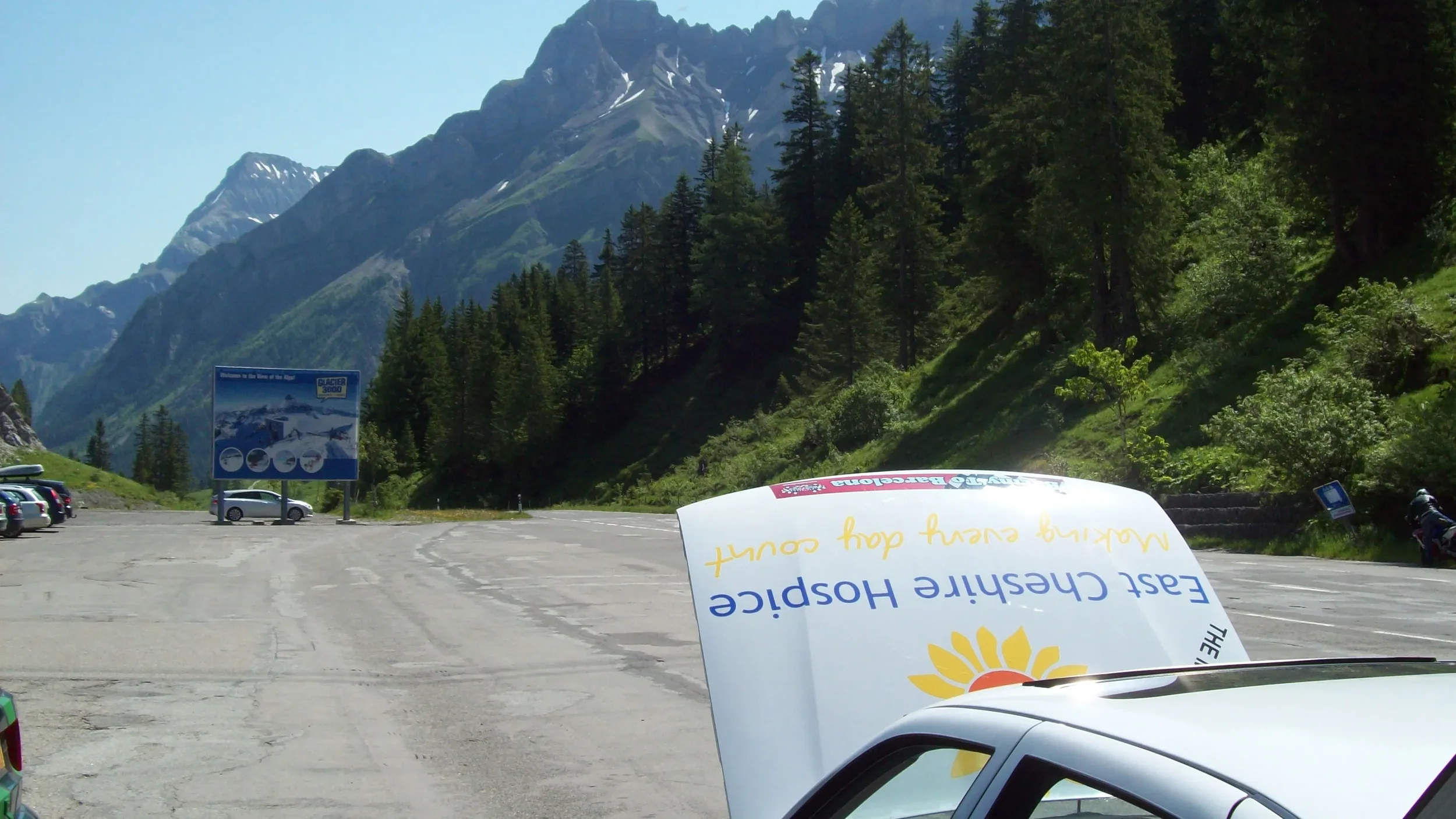 A parking lot in a mountain area with lush green trees, tall mountains in the background, and a blue informational sign. There is a partially visible car in the foreground with a sign on its hood that has the logo and text indicating it's part of a t
