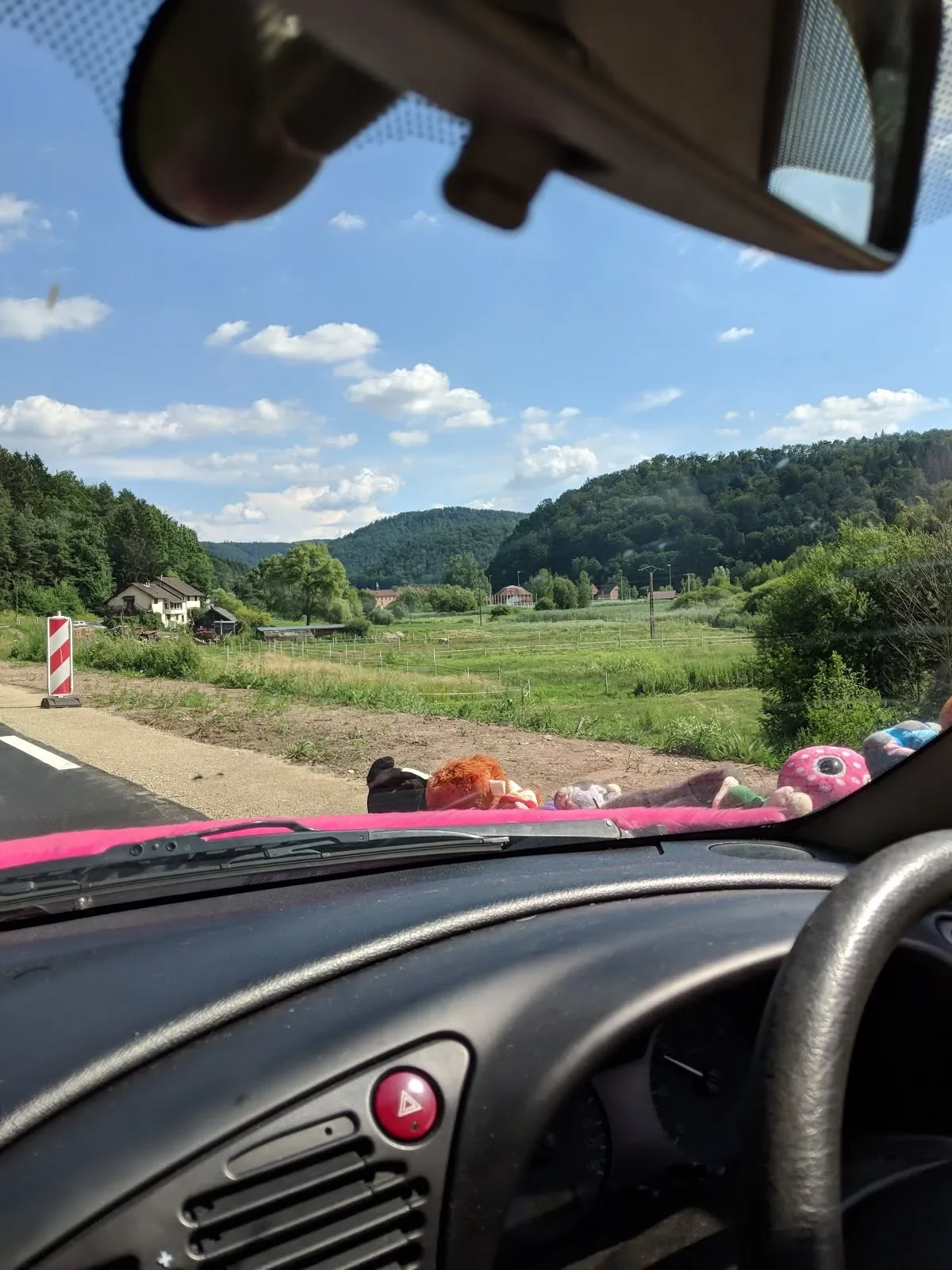 View from inside a car showing a rural landscape with green fields, trees, and hills under a blue sky with scattered clouds. Plush toys are placed on the dashboard.