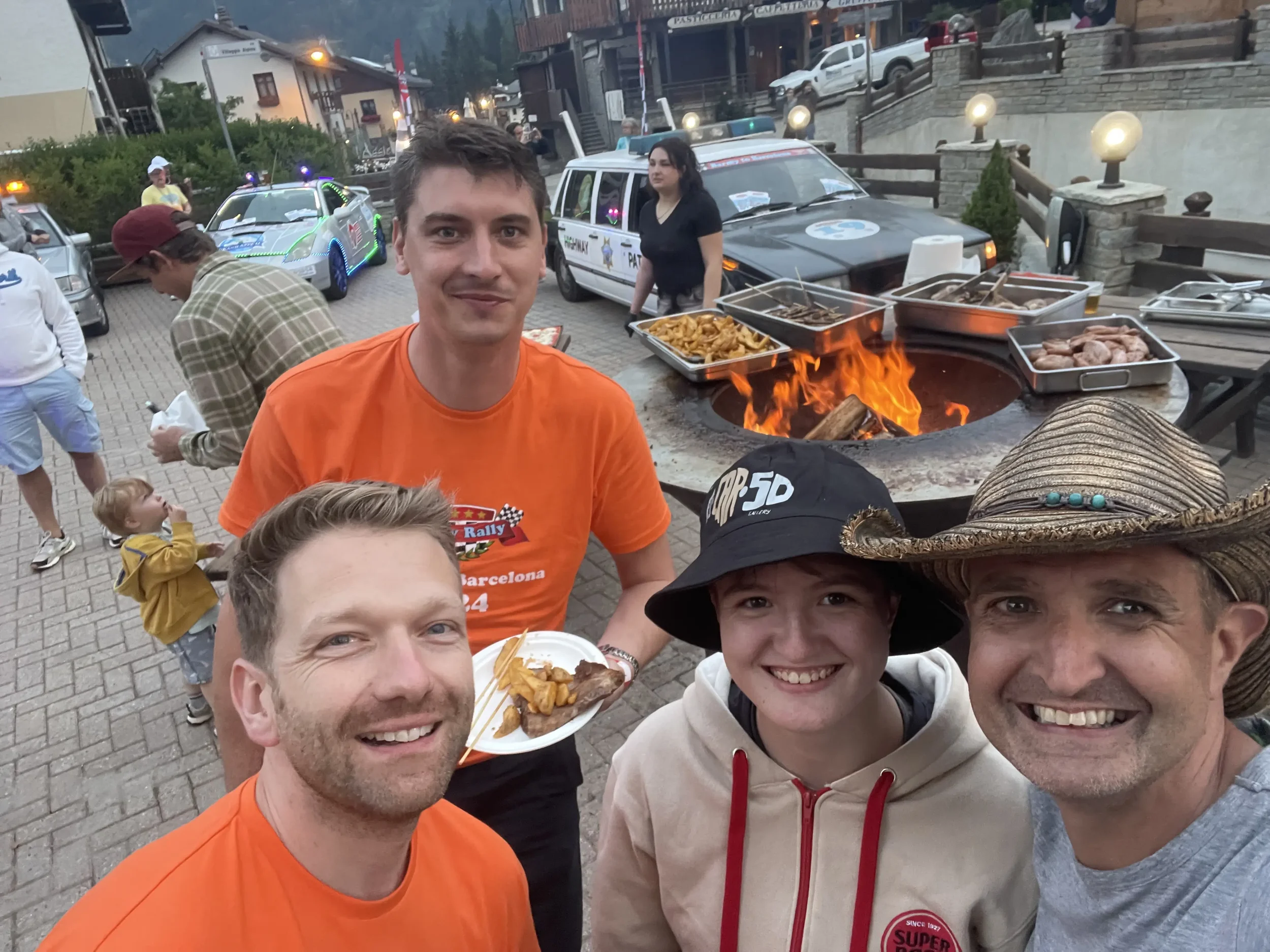 Group of five people taking a selfie at an outdoor event with a fire pit behind them. There are food trays, police cars, and a small child in the background.