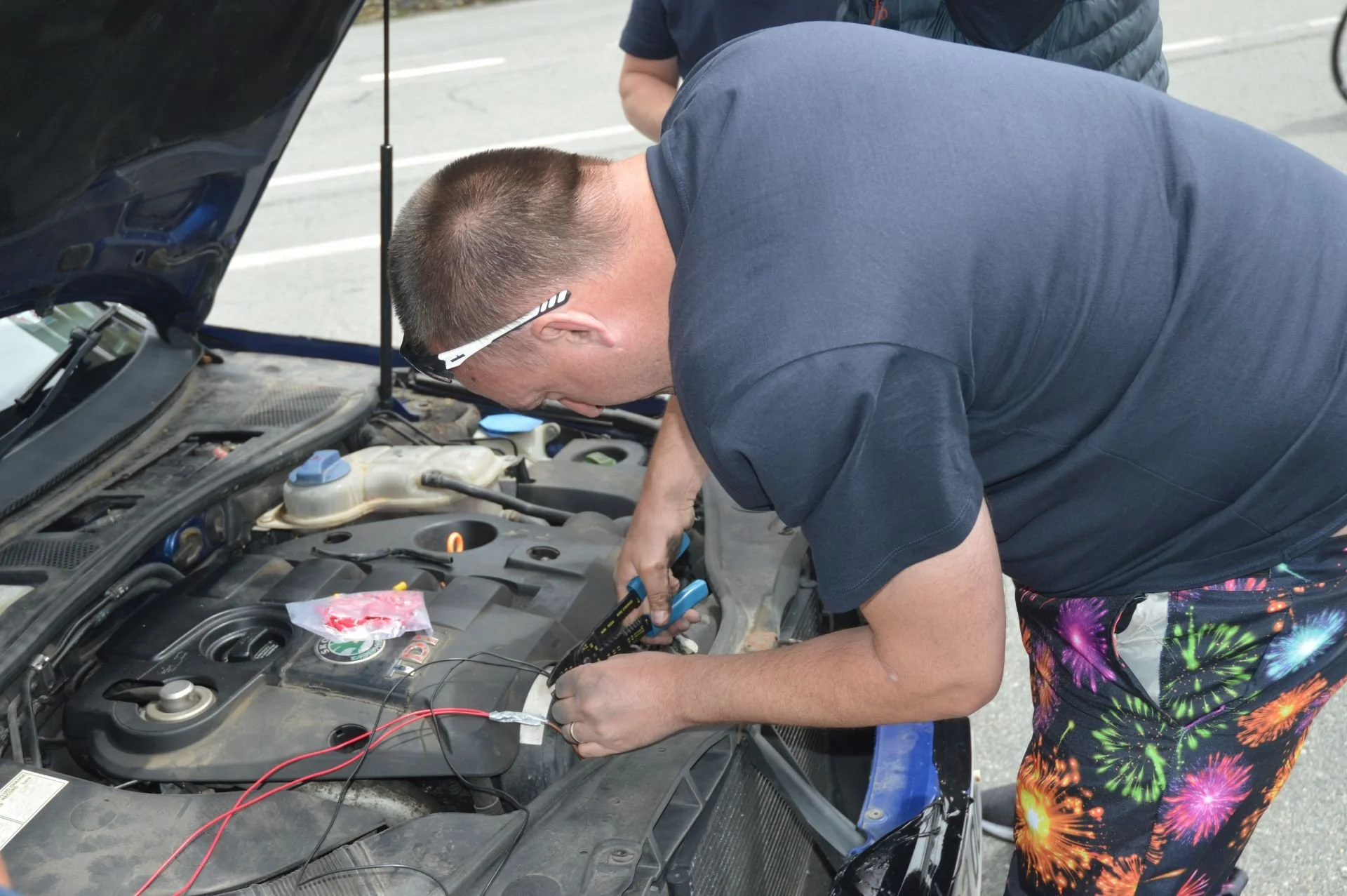 A man wearing sunglasses and colorful fireworks-patterned shorts working under the hood of a car in a parking lot, using electrical testing tools.