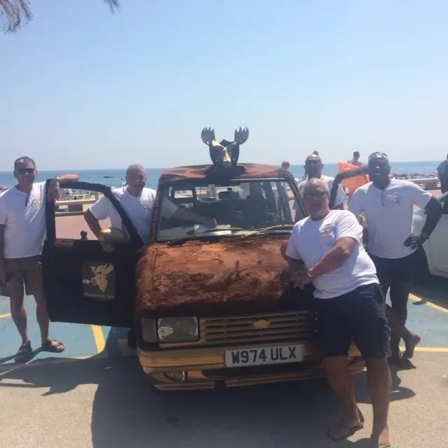 Group of people posing around a vehicle with a moose head on its roof at the beach parking lot.