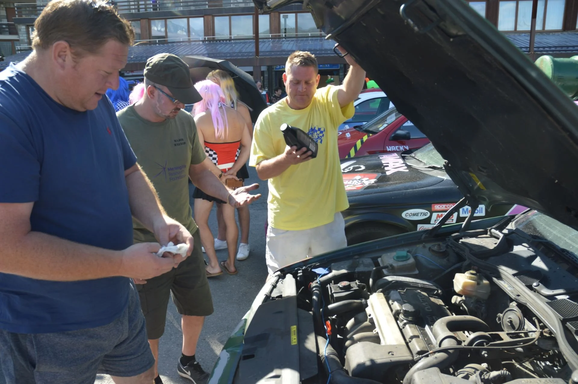 Group of people gathered around a car with its hood open, inspecting the engine at an outdoor event.