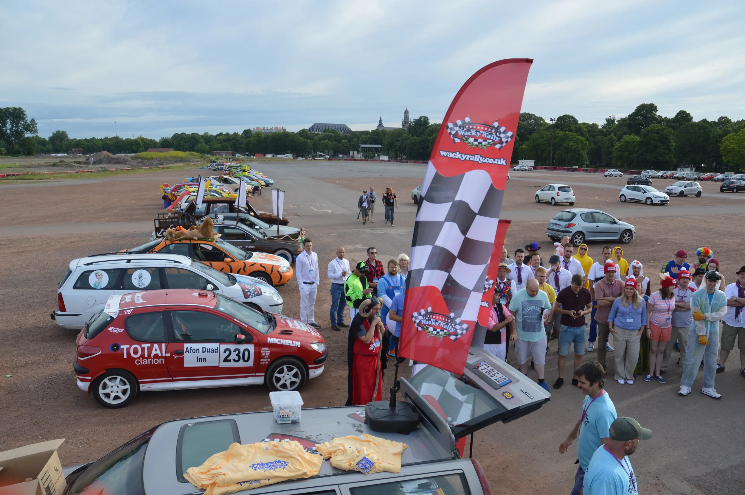 Car racing event with a line of decorated cars, people in costumes, and a large red flag with a checkered flag and logo that reads Wacky Races. Participants and spectators are gathered near the cars on a dirt lot, with some people taking photos, and 