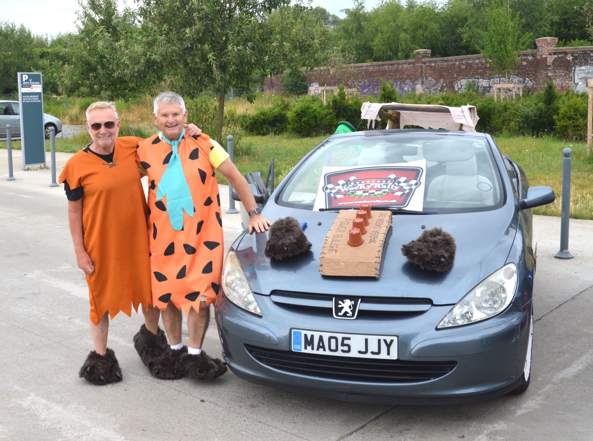 Two men dressed in Fred Flintstone costumes standing next to a decorated car with a sign on the hood that reads "Race Car" and two plush animal heads on the hood, in a parking lot with greenery and a brick wall in the background.