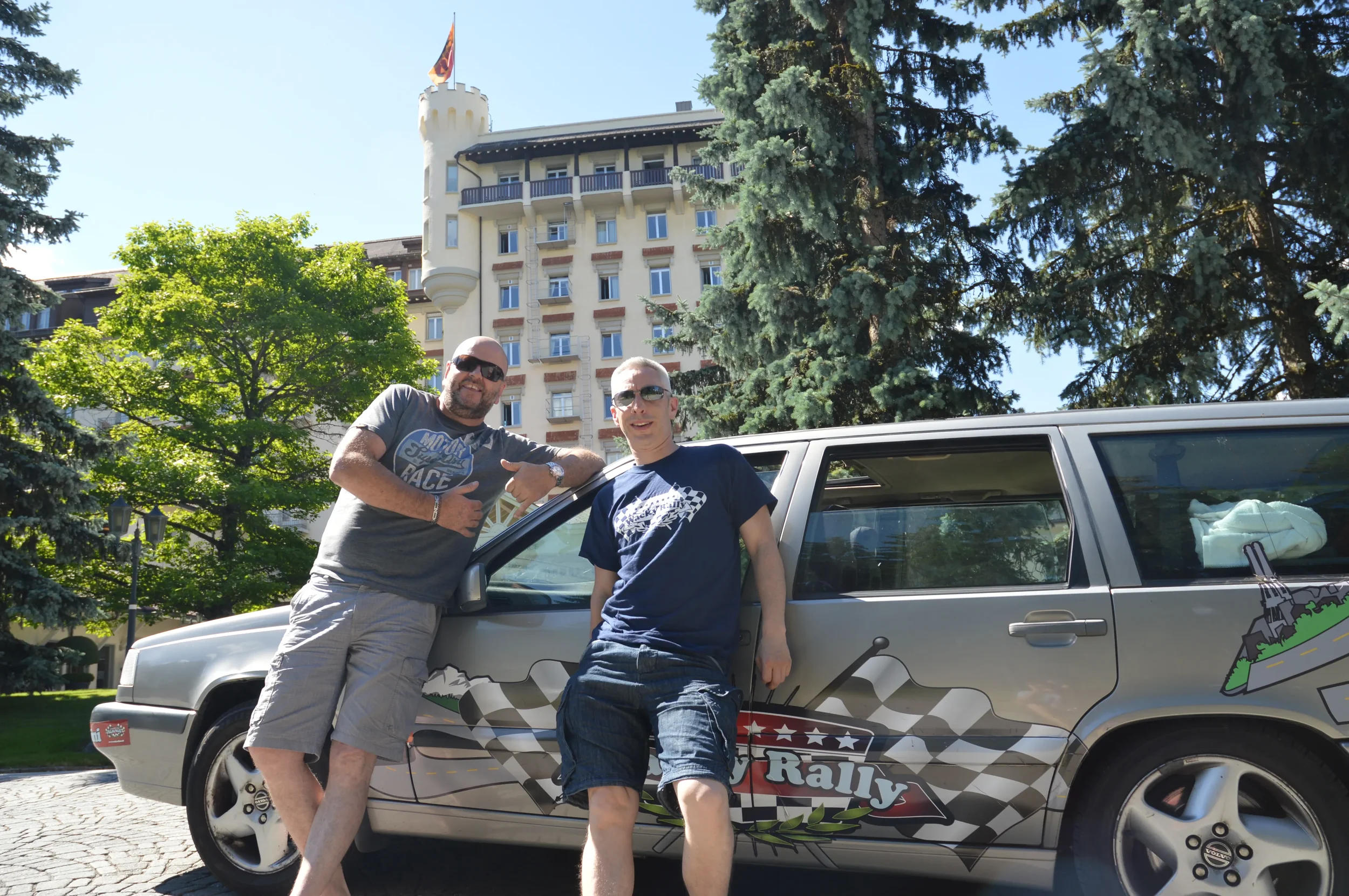 Two men posing next to a silver rally car with a design of checkered flags, mountains, and a motorcycle. One man wearing sunglasses, gray t-shirt, and khaki shorts. The other man wearing sunglasses, navy t-shirt, and denim shorts. Large trees and a b