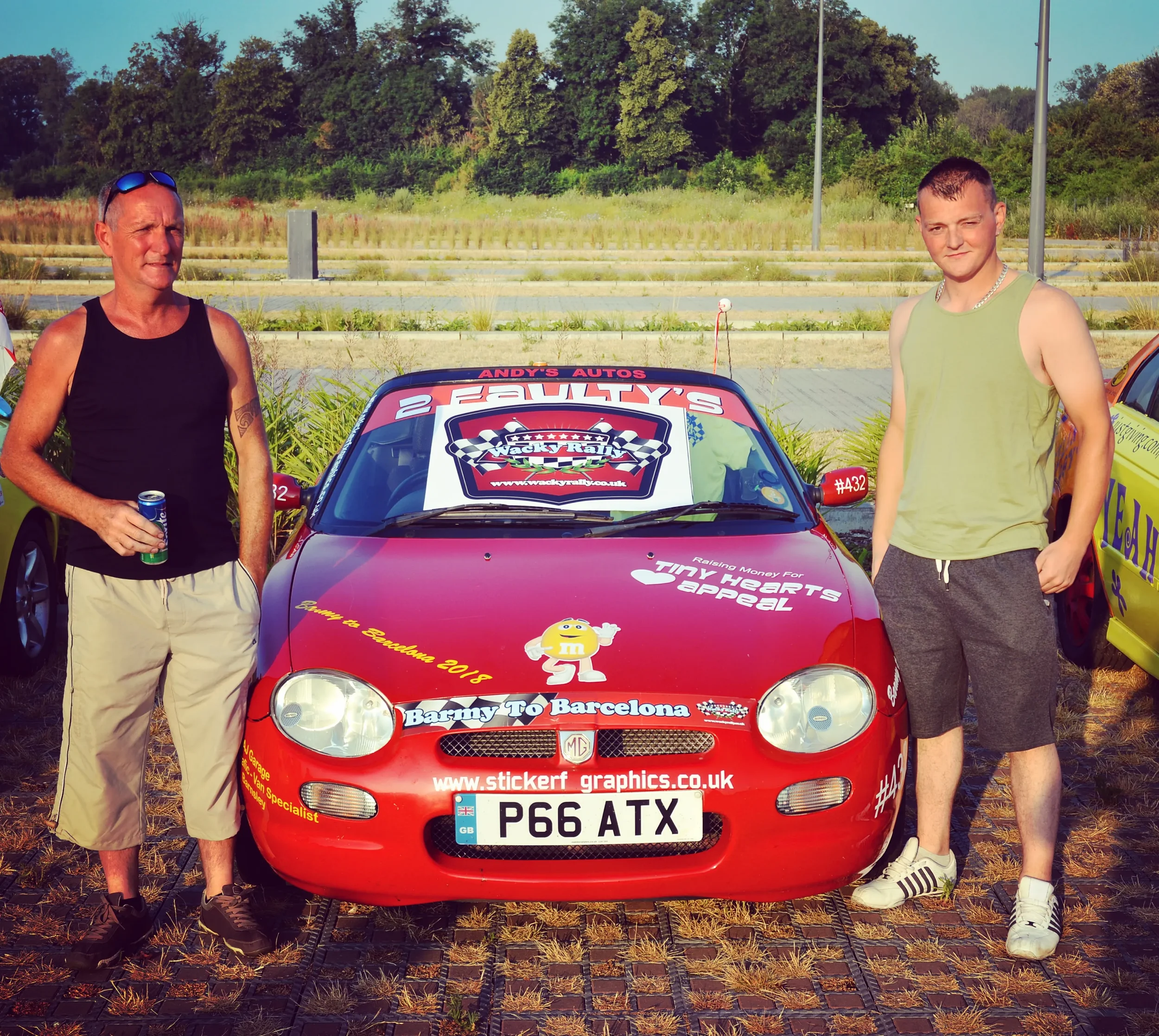 Two men standing beside a red racing car with various sponsor stickers at an outdoor event.