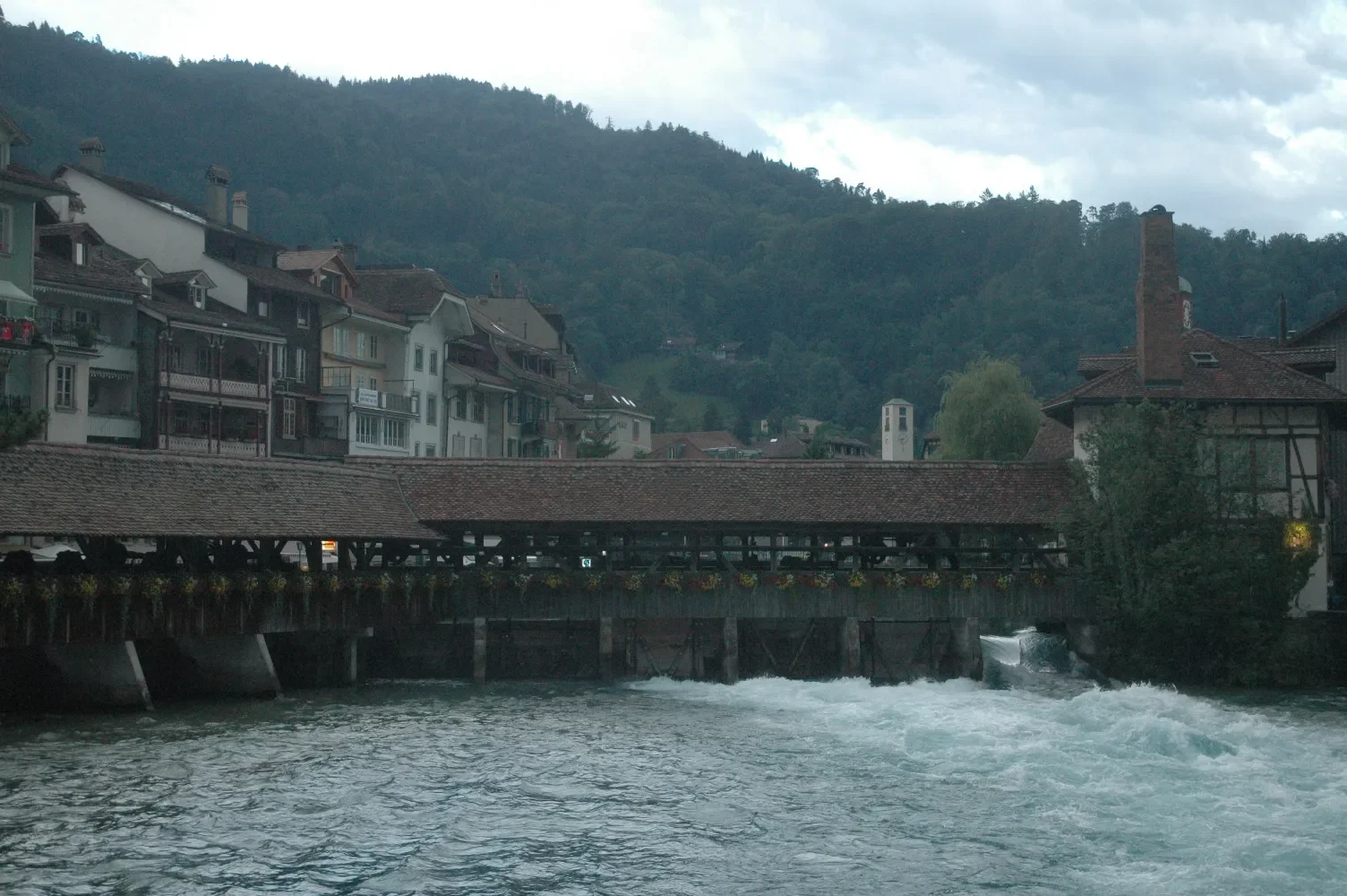 A riverside town with tightly packed buildings on a hillside, a wooden covered bridge over the water, mountains in the background, and a cloudy sky.