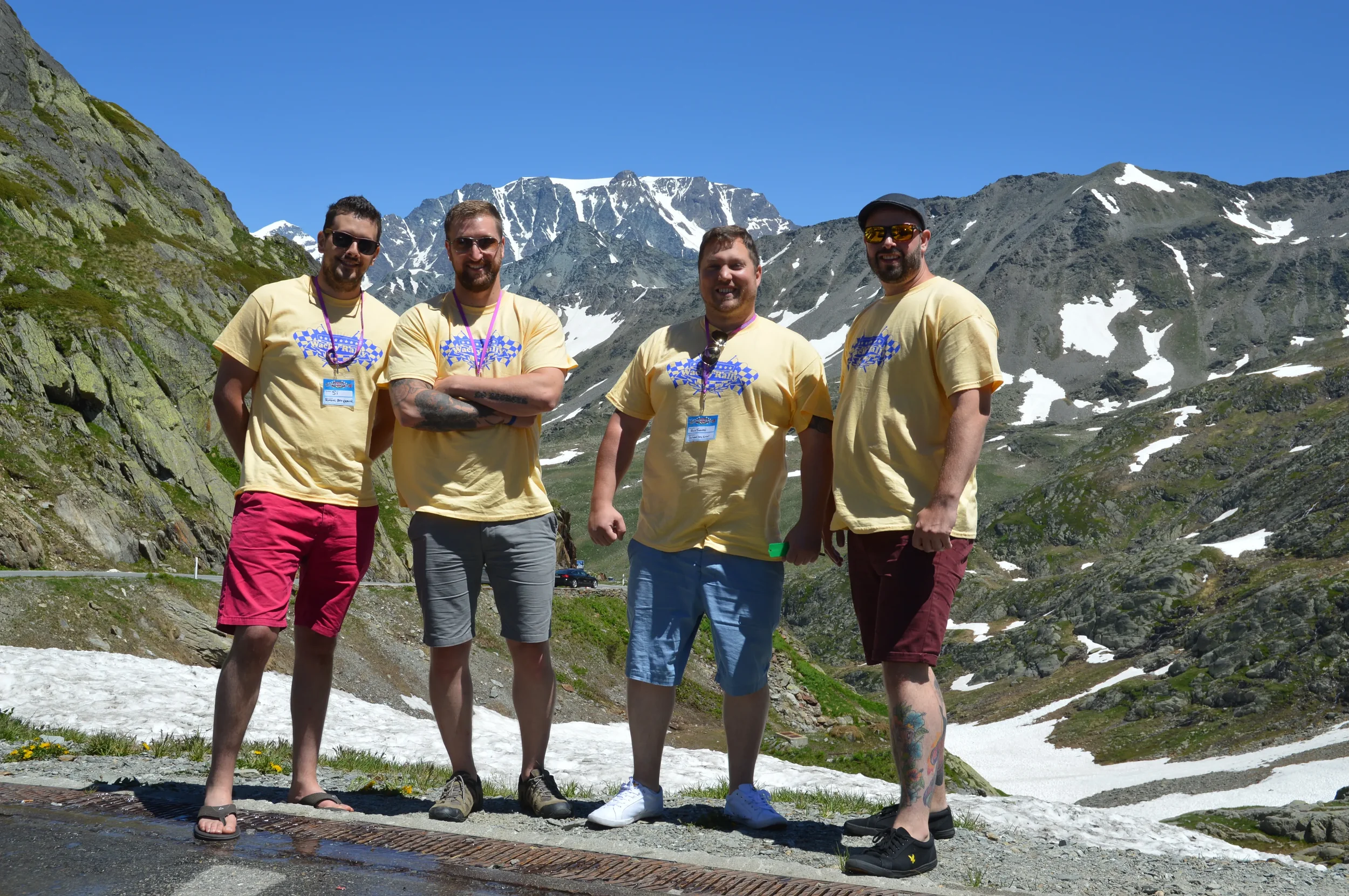 Four men standing together outdoors in a mountainous area with snow patches and rocky terrain, all wearing yellow T-shirts with a blue graphic and sunglasses, smiling at the camera.