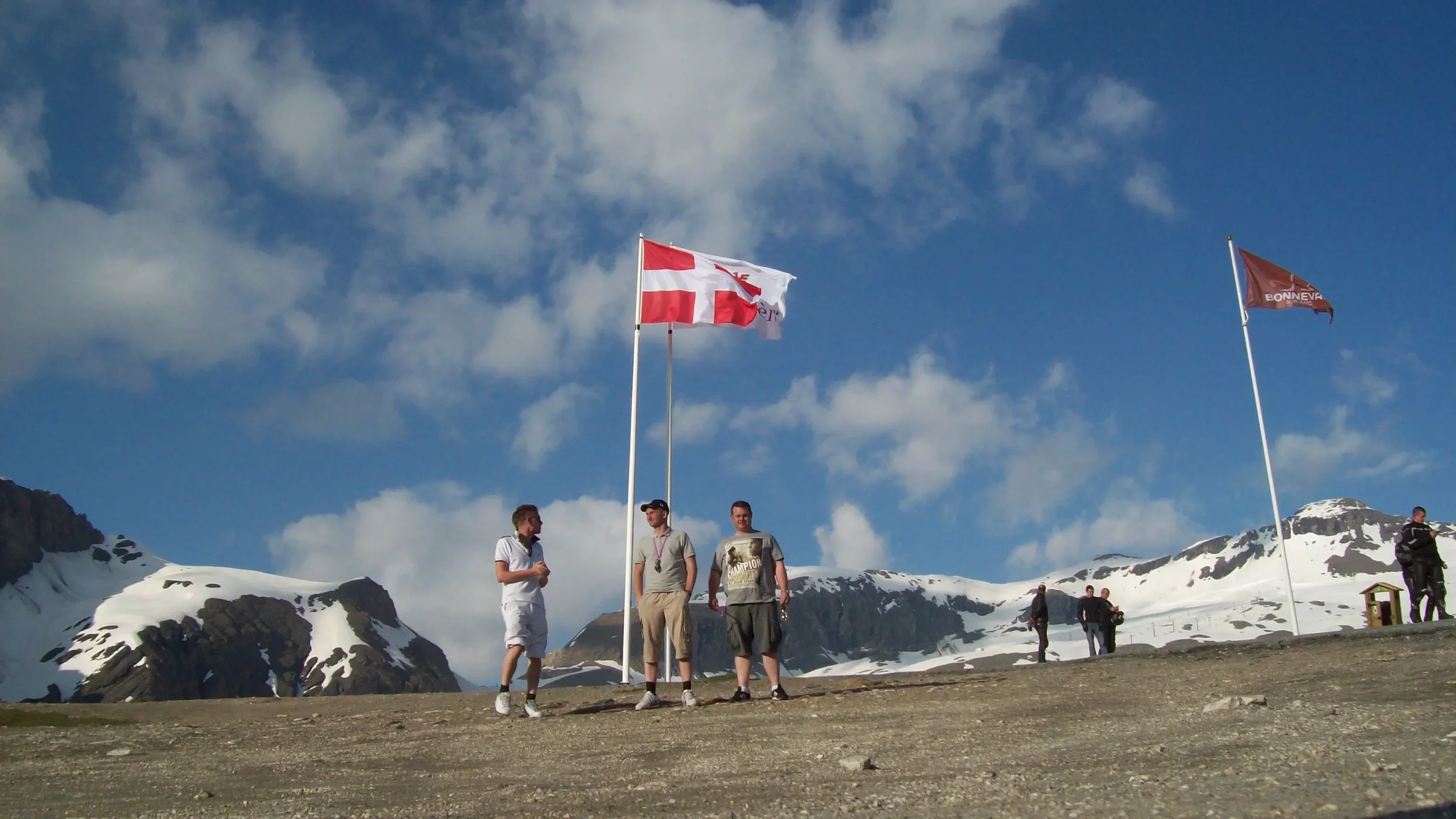 Three men standing near a flagpole with a Danish flag and another flag, with snow-covered mountains and a partly cloudy sky in the background.