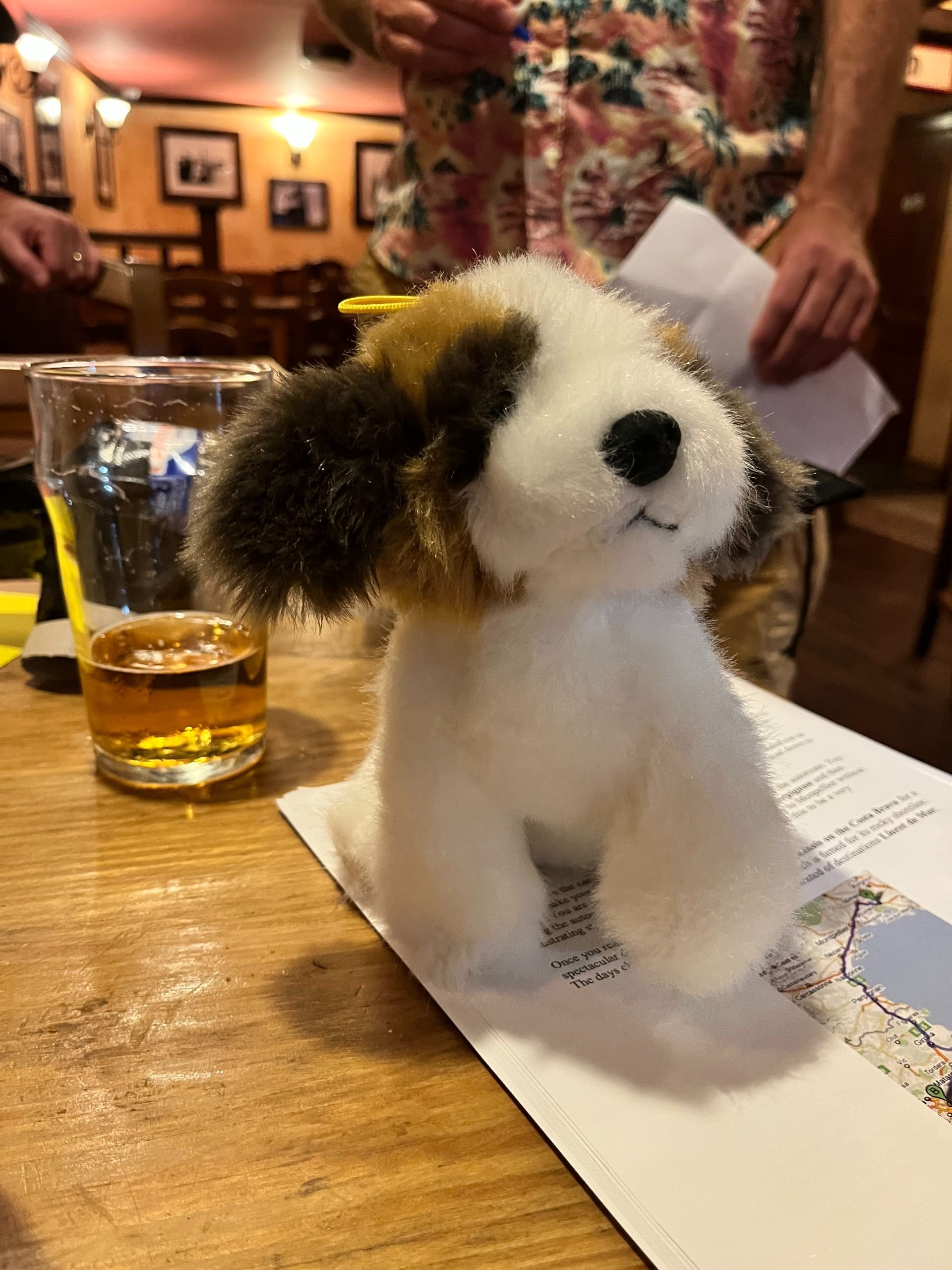 A plush toy dog is placed on a table in a restaurant, with a glass of beer and a newspaper nearby. People are standing in the background.