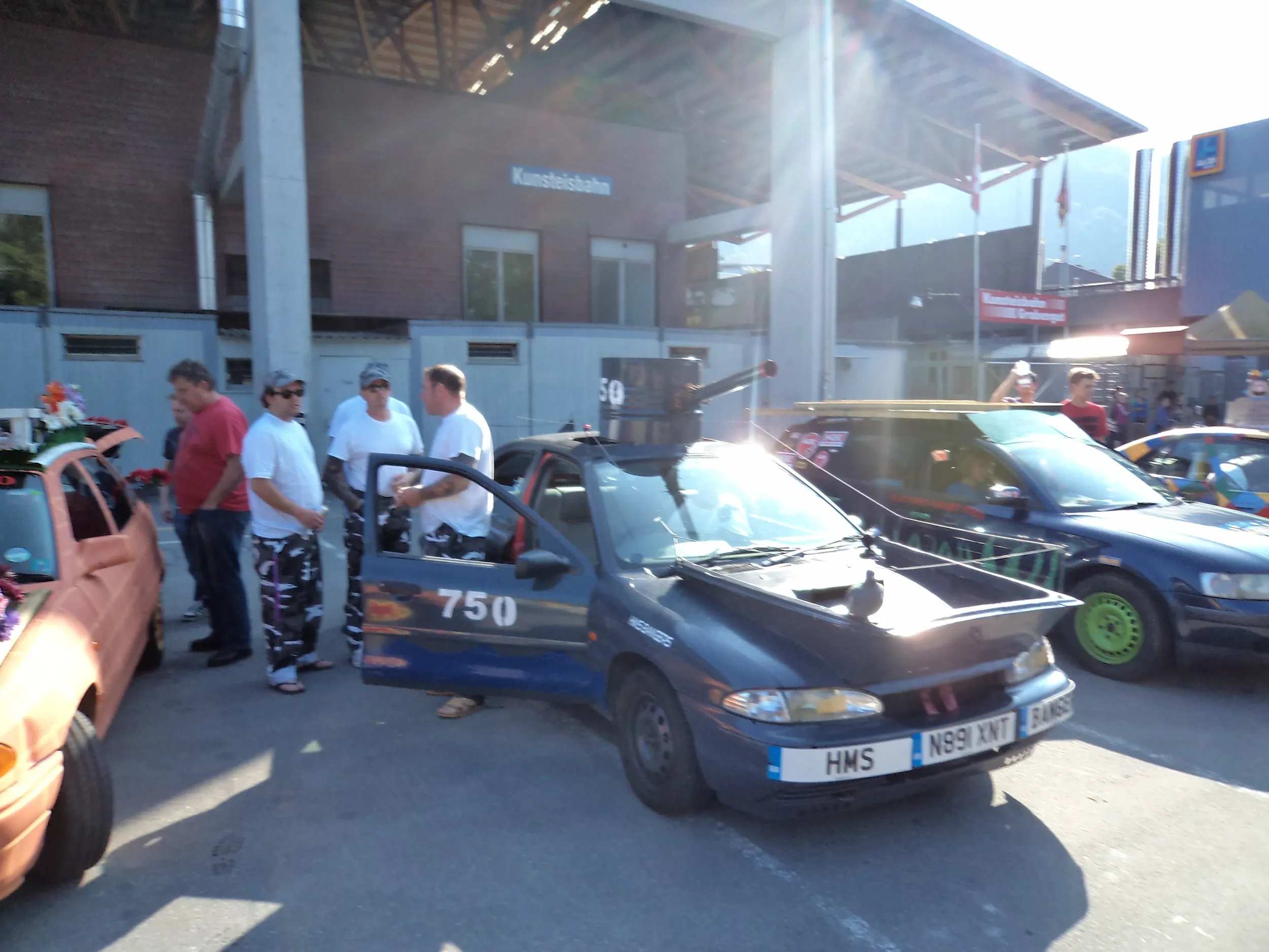 Group of people standing around a small black car with a mounted cannon on top, parked beside other colorful cars, at an outdoor event under sunlight.