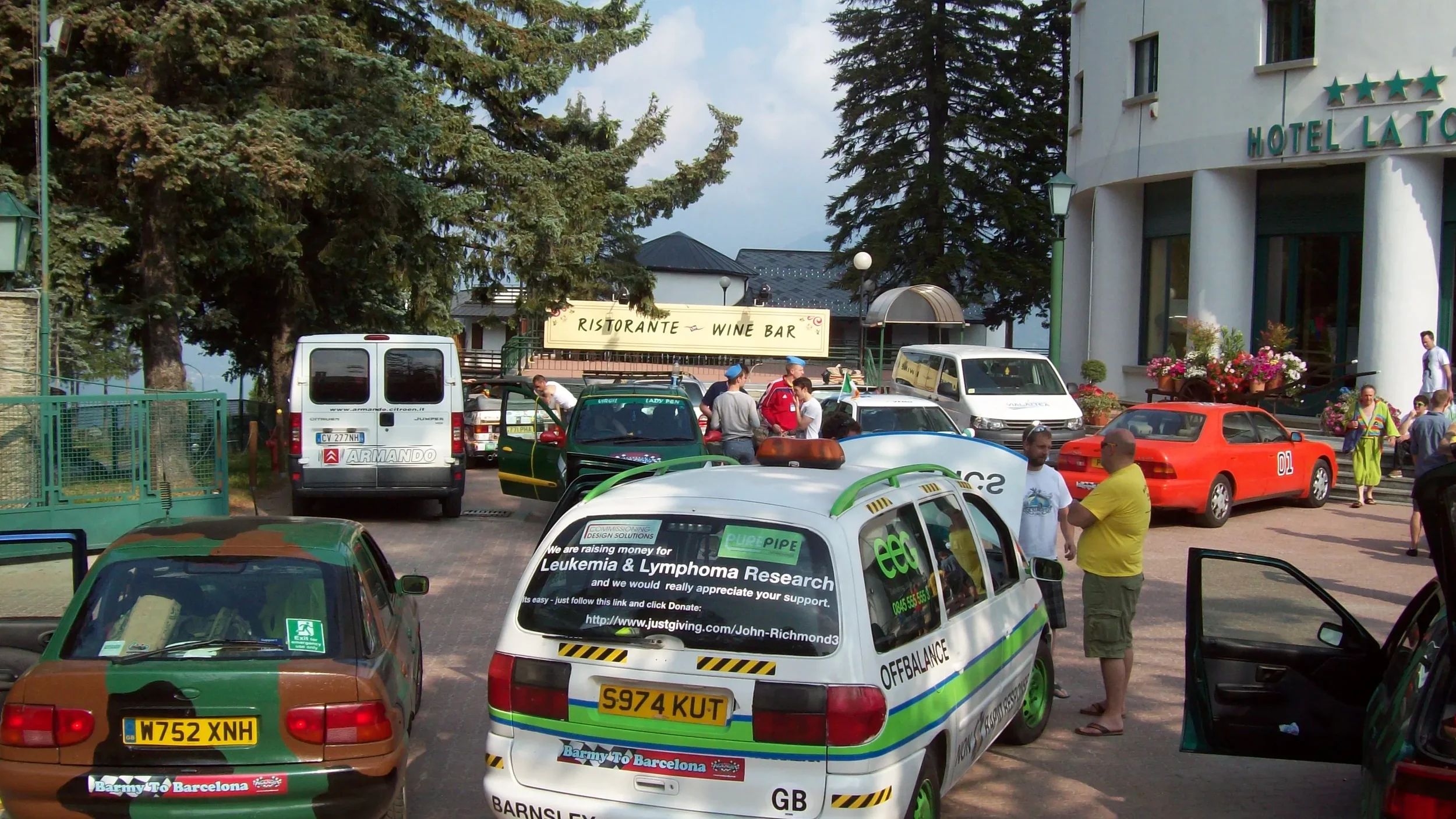 Parking lot filled with cars and people outside a hotel with a sign that says 'Ristorante - Wine Bar'; green trees in the background.