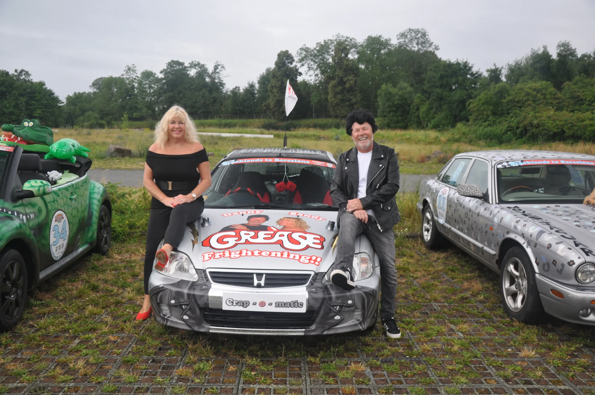 Two smiling people, a woman and a man, standing next to a decorated car on a cloudy day, with other decorated cars and trees in the background.