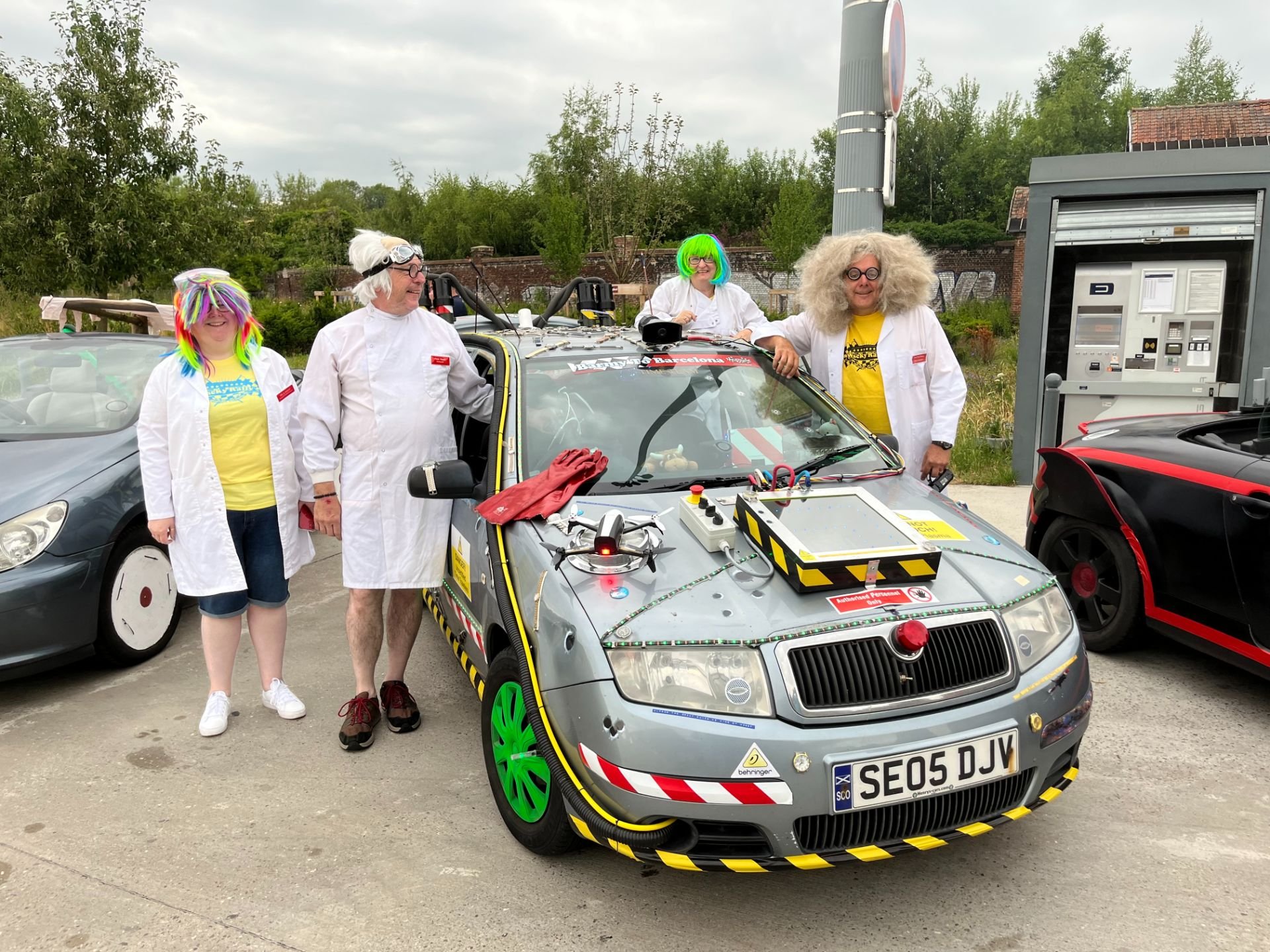 Four people dressed as scientists with colorful wigs and lab coats standing around a small, modified car with scientific equipment on it, outdoors near a parking area.