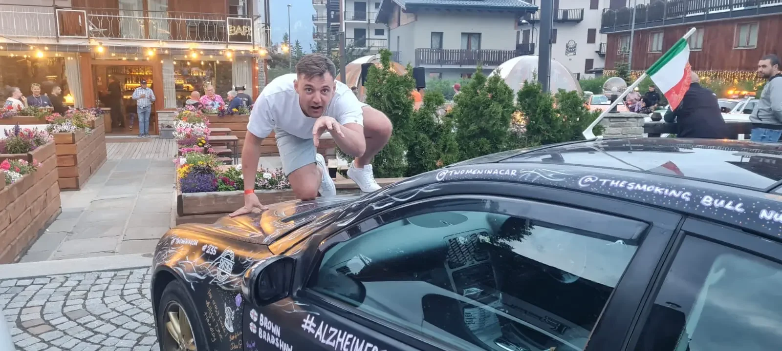 A man crouches on a black race car with colorful lightning bolt designs, pointing towards the camera. The car has social media handles and a hashtag '#ALZHEIMER'. In the background, people are standing outside a restaurant with outdoor seating and fl
