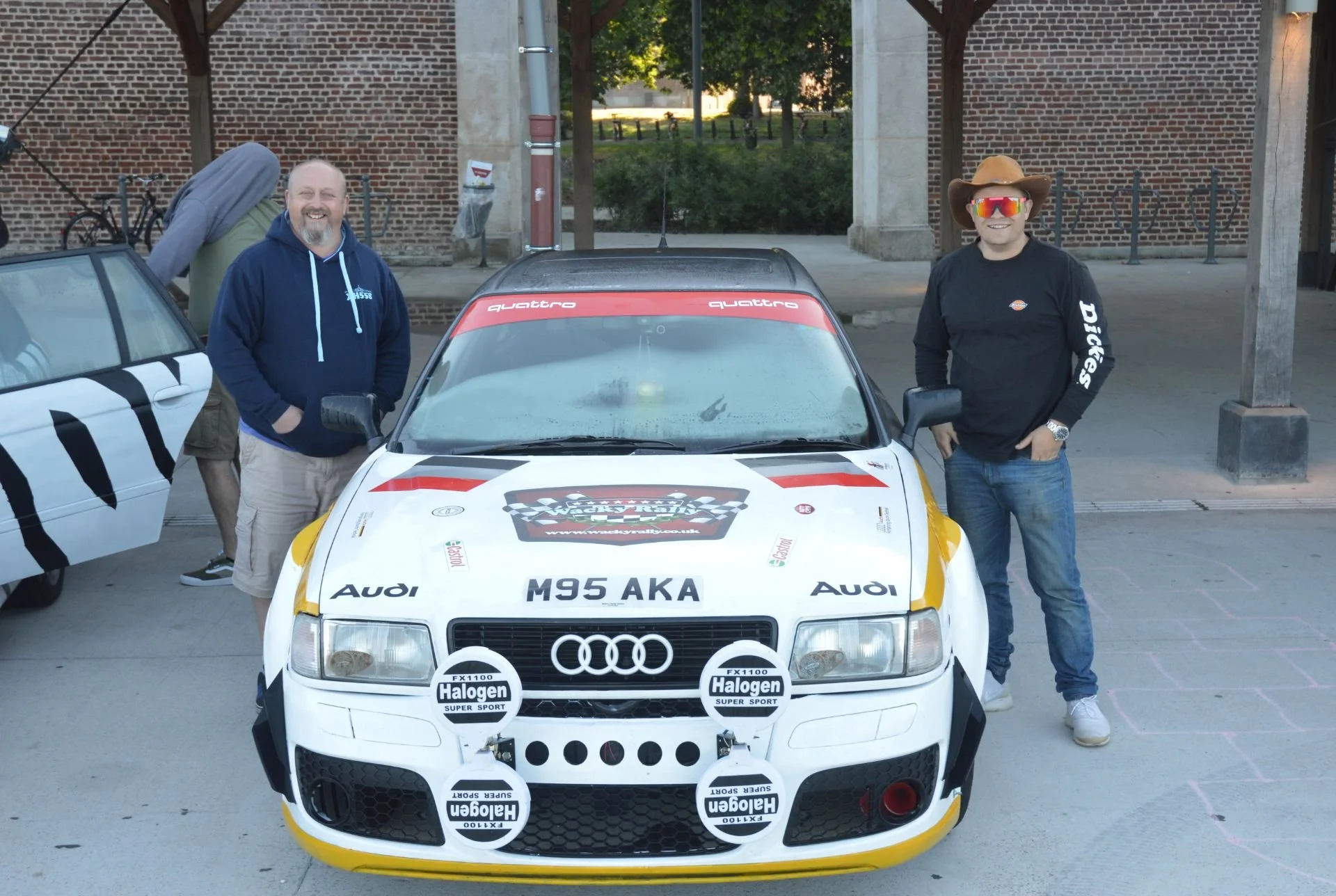 Two men standing next to a white Audi racing car with a checkered flag logo, in front of brick buildings. One man wearing a blue hoodie, the other wearing a cowboy hat, sunglasses, and a black long-sleeve shirt.