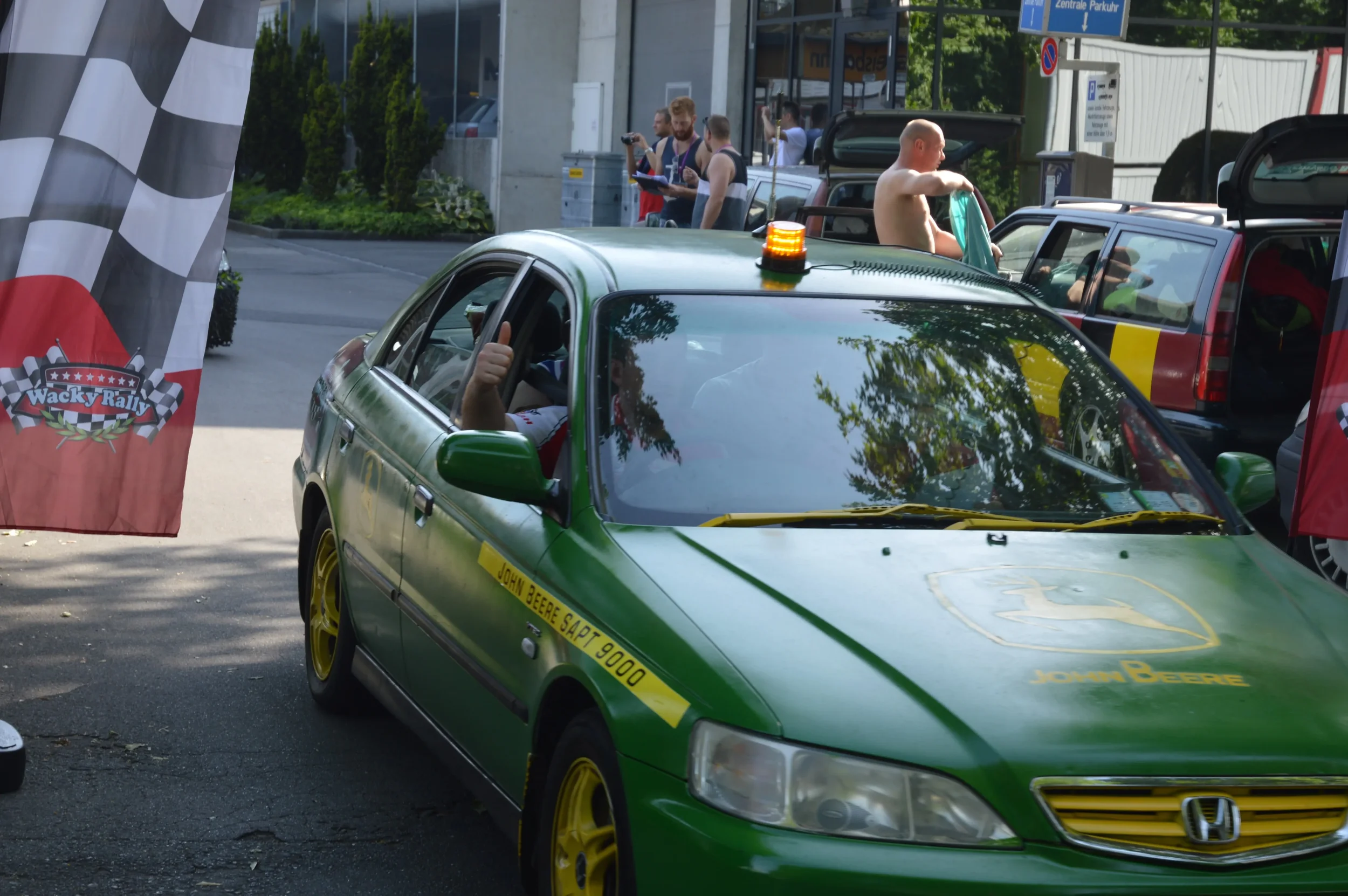 A green race car with yellow wheels and John Deere branding is parked at a rally event. A person inside is giving a thumbs up, while several people in the background watch and record the event.