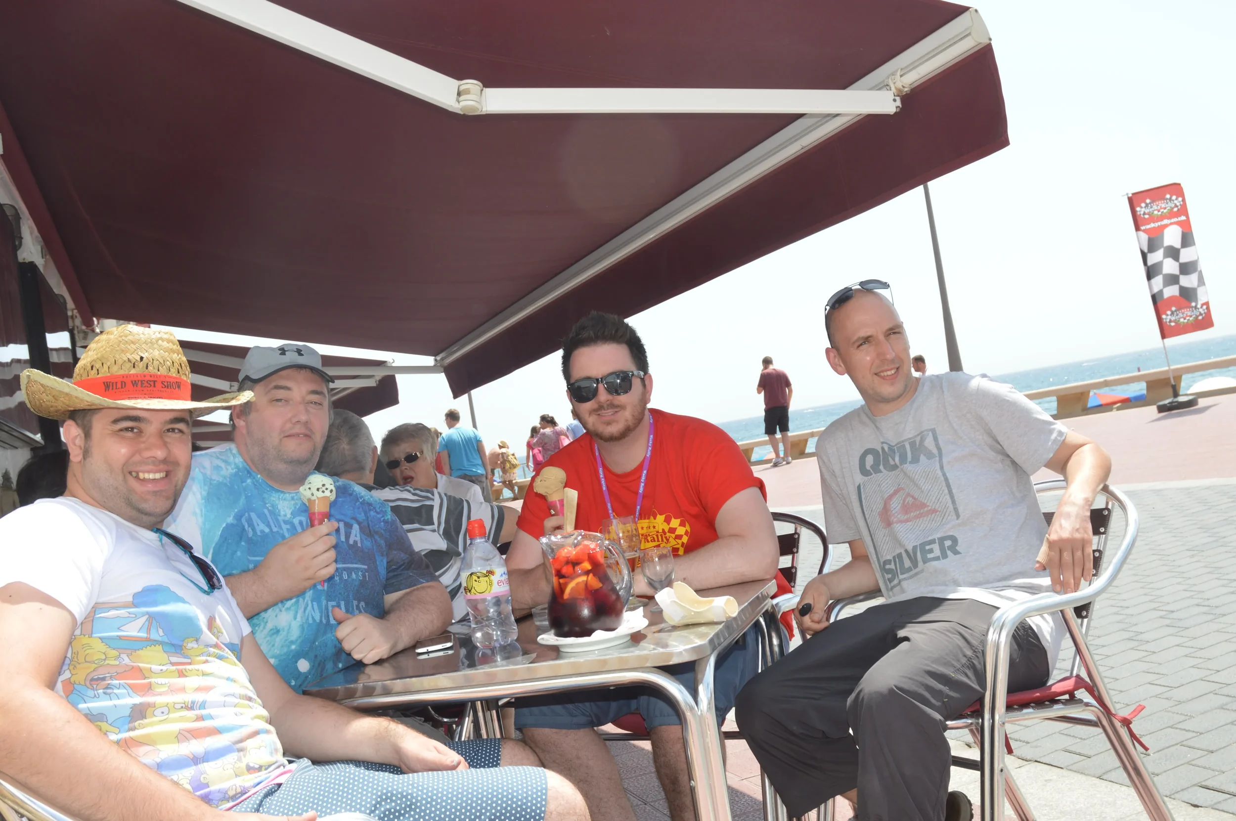 Four men sitting at a table outdoors near the water, enjoying ice cream, drinks, and food in a sunny setting with a red canopy overhead.