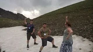 Three people standing outdoors on rocky terrain, with two of them posing with legs apart and hands raised, while the third looks on. A cloudy sky is in the background.