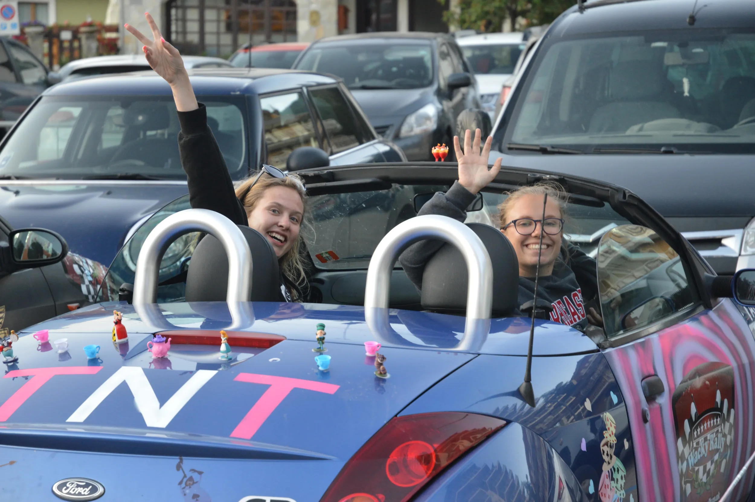 Two young women in a decorated blue convertible car are waving and smiling at the camera, with cars parked on the street behind them.