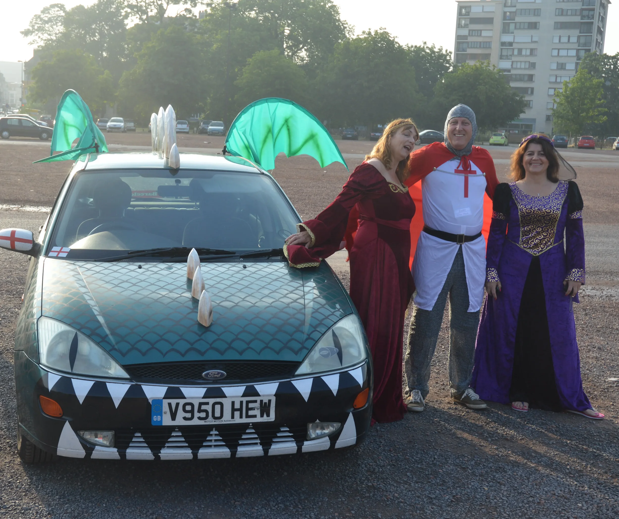 People dressed in medieval costumes standing next to a decorated car with dragon wings, shields, and a dragon face on the front, in an outdoor parking lot.