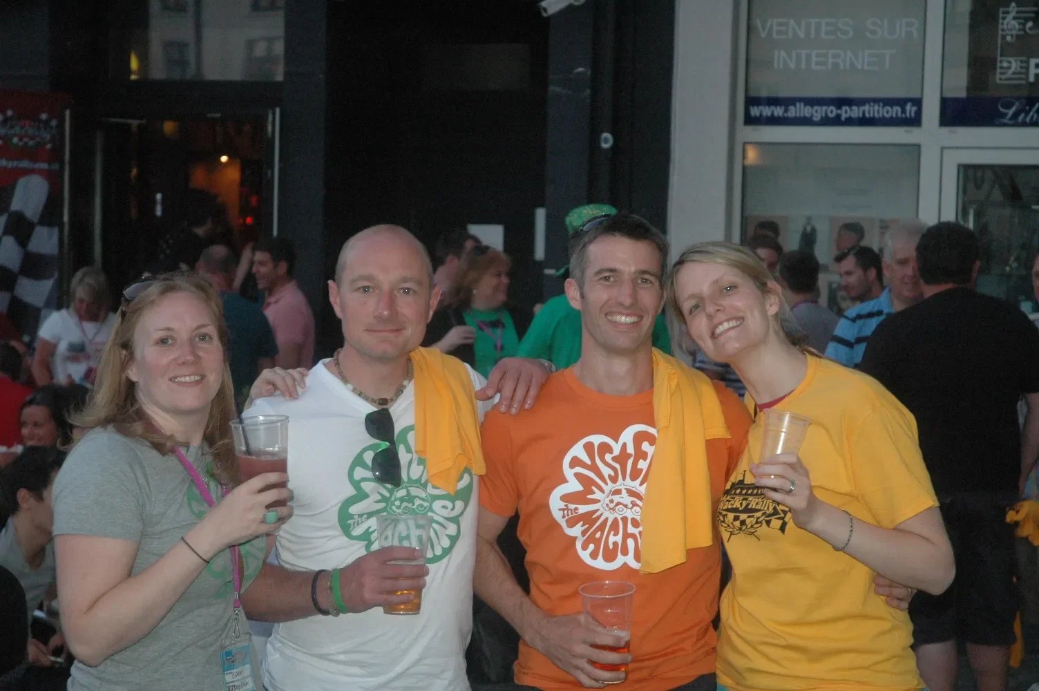 Four people at a social event, smiling and holding drinks, with a crowd in the background.