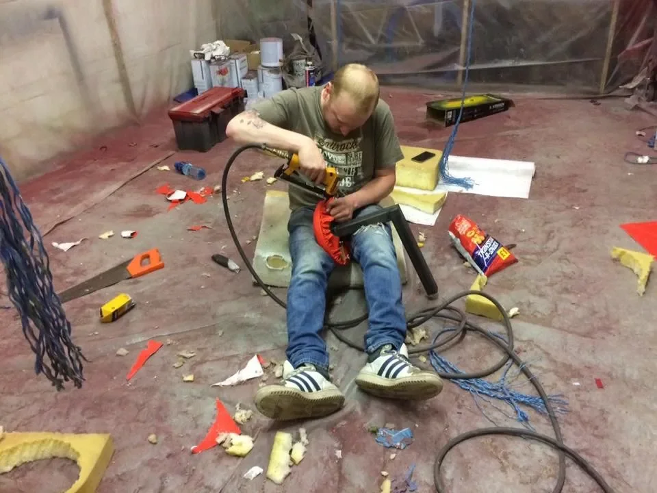 A man is sitting on a small revolved seat in a construction or workshop setting, working with a power drill on a red circular object, surrounded by scattered tools, debris, and insulation material.