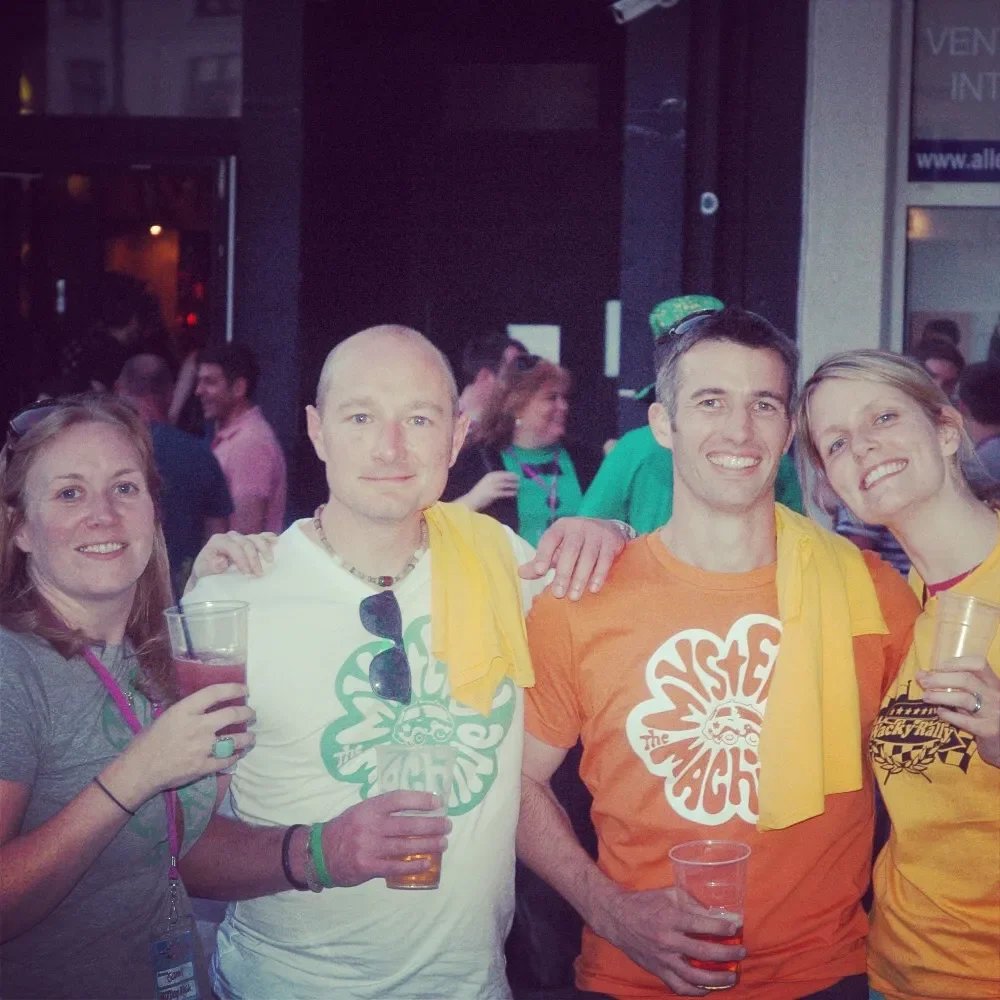 Four friends at a party, holding drinks and smiling. They are wearing colorful t-shirts and have yellow towels draped over their shoulders.
