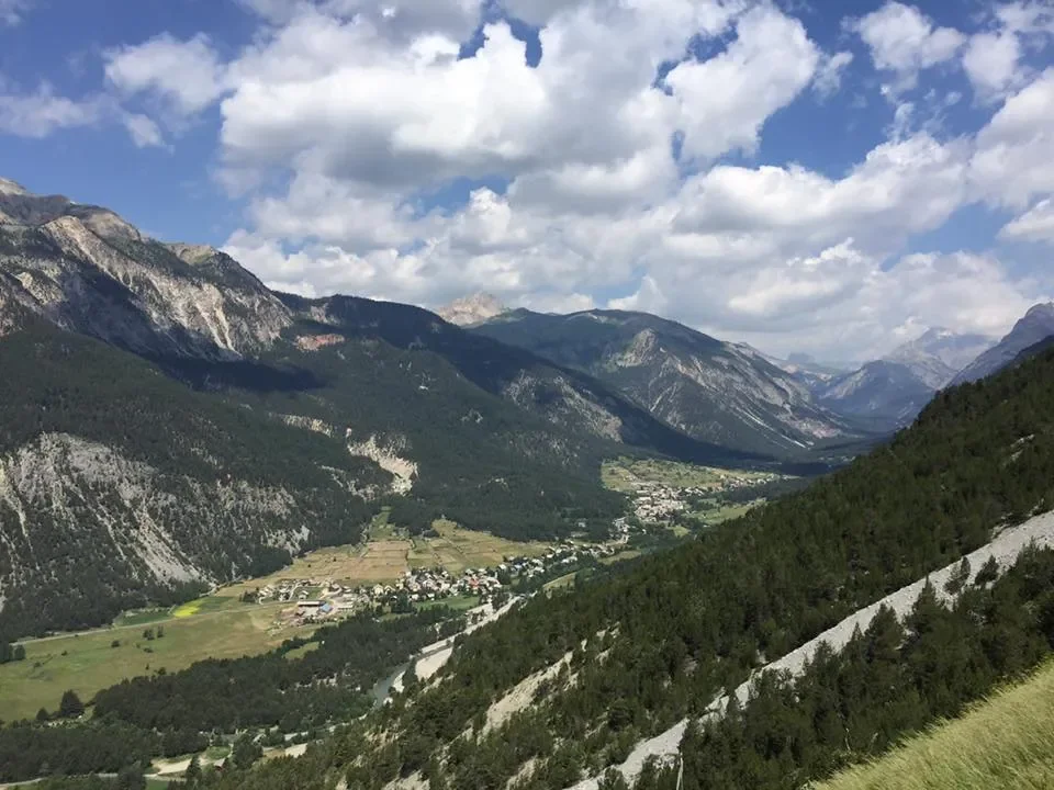 Scenic view of a mountain valley with green fields, a small village, and forested slopes under a partly cloudy sky.