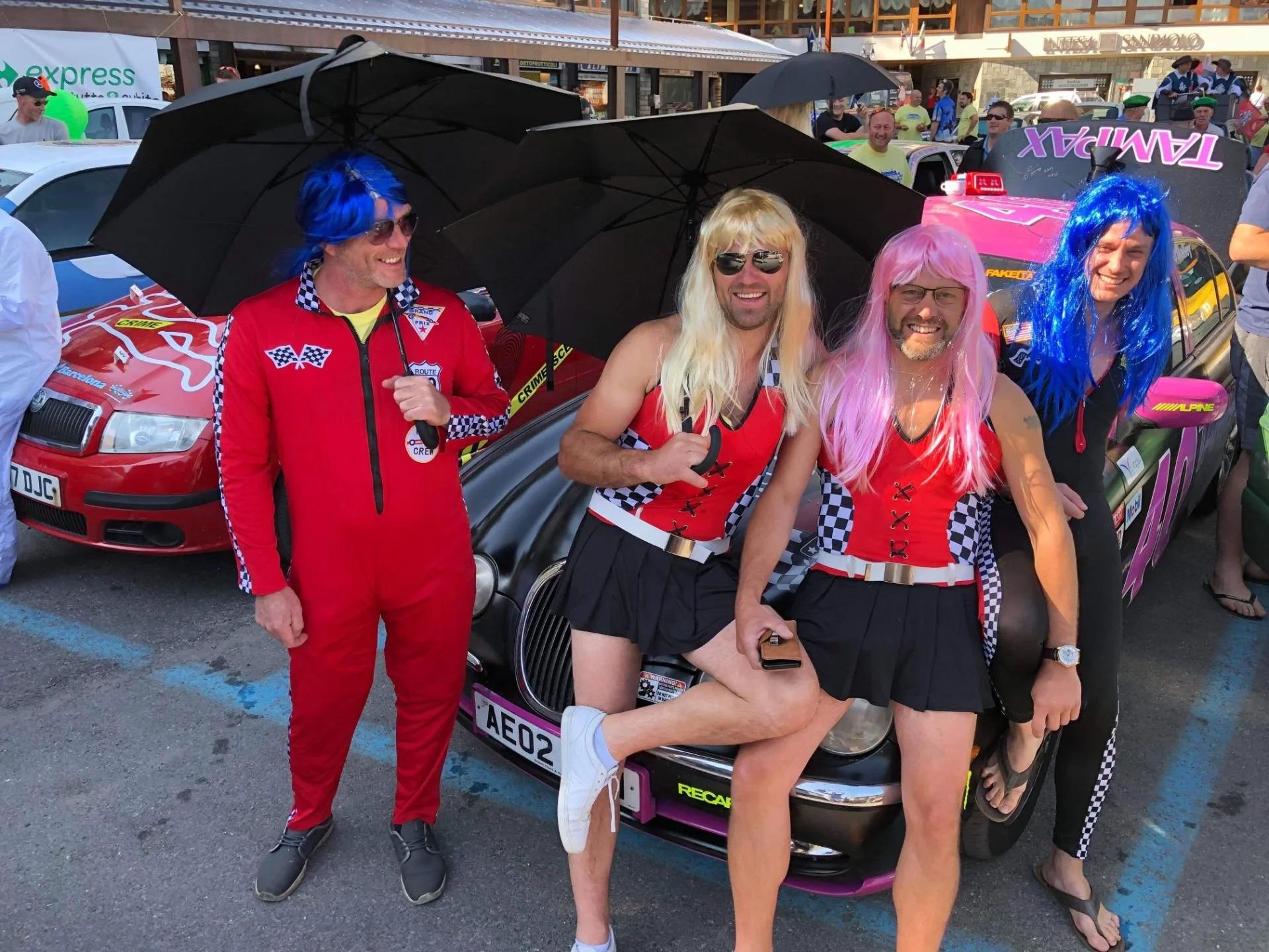 Four men in racing uniforms with wigs, sunglasses, and umbrellas posing in front of a race car at a racing event. Three of them are wearing colorful wigs and racing outfits, while one is dressed in a red jumpsuit.