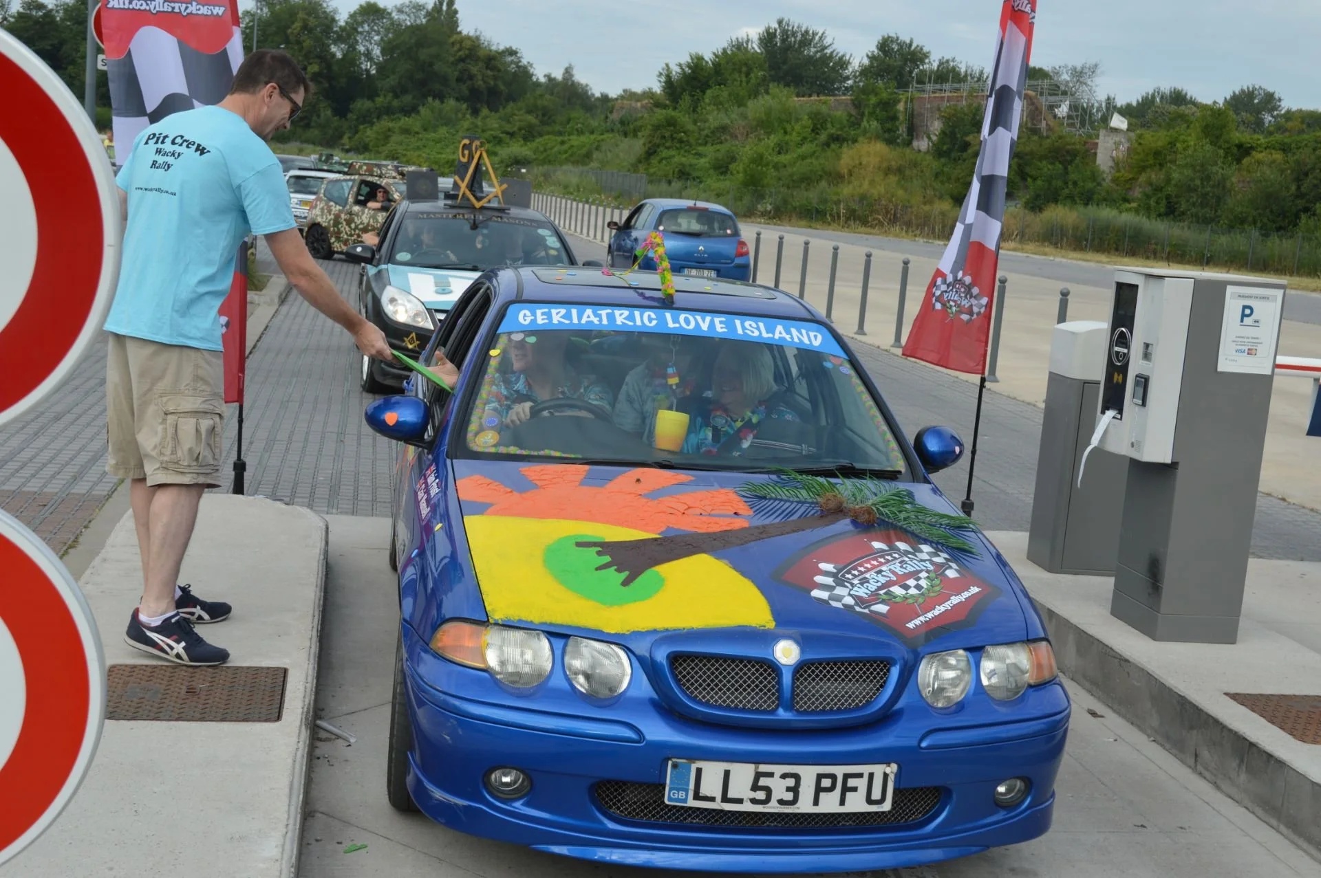 A decorated blue car with a large sign on the windshield reading 'Geriatric Love Island' parked at a toll booth. Inside, people are dressed in colorful outfits. A man wearing a light blue shirt labeled 'Pit Crew Wacky Rally' is handing a green object