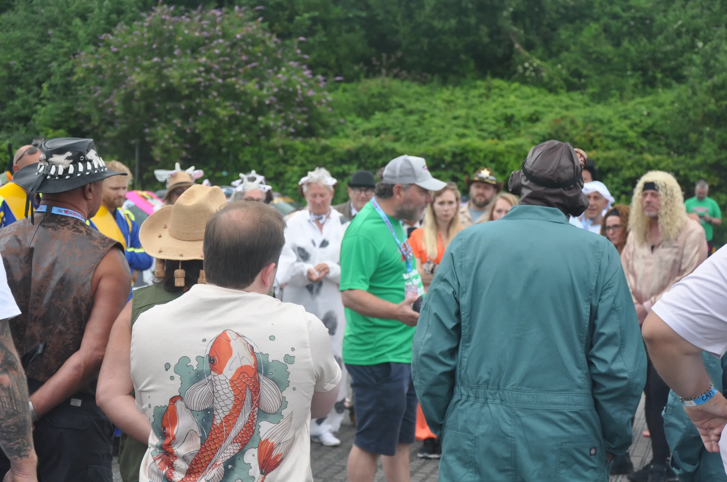 A group of people gathered outdoors in a park with trees and greenery in the background. Some are dressed in costumes, and one person in the foreground is wearing a shirt with a large koi fish illustration.