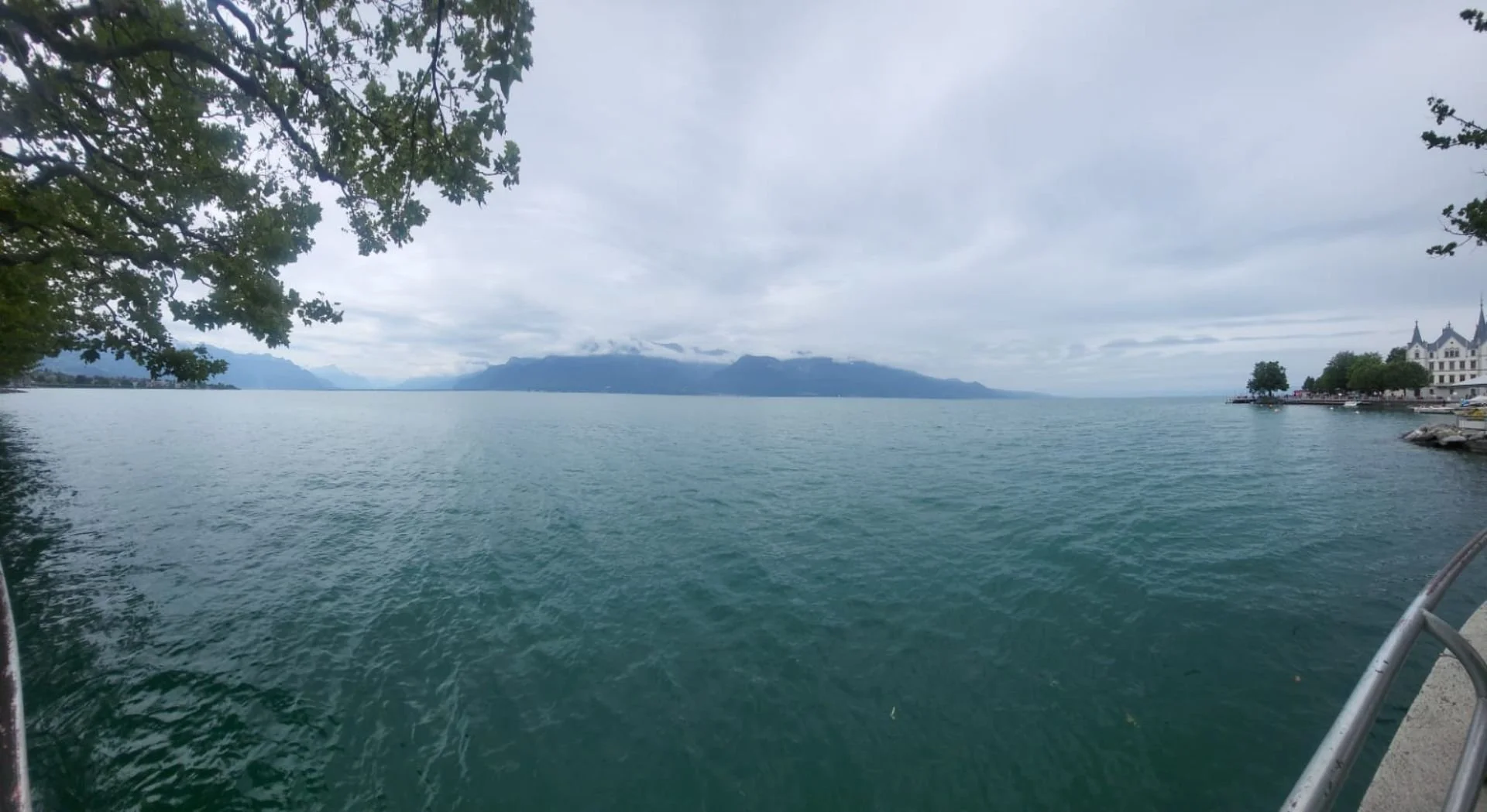 View of a large lake with mountains in the distance, trees along the shoreline, and a cloudy sky.
