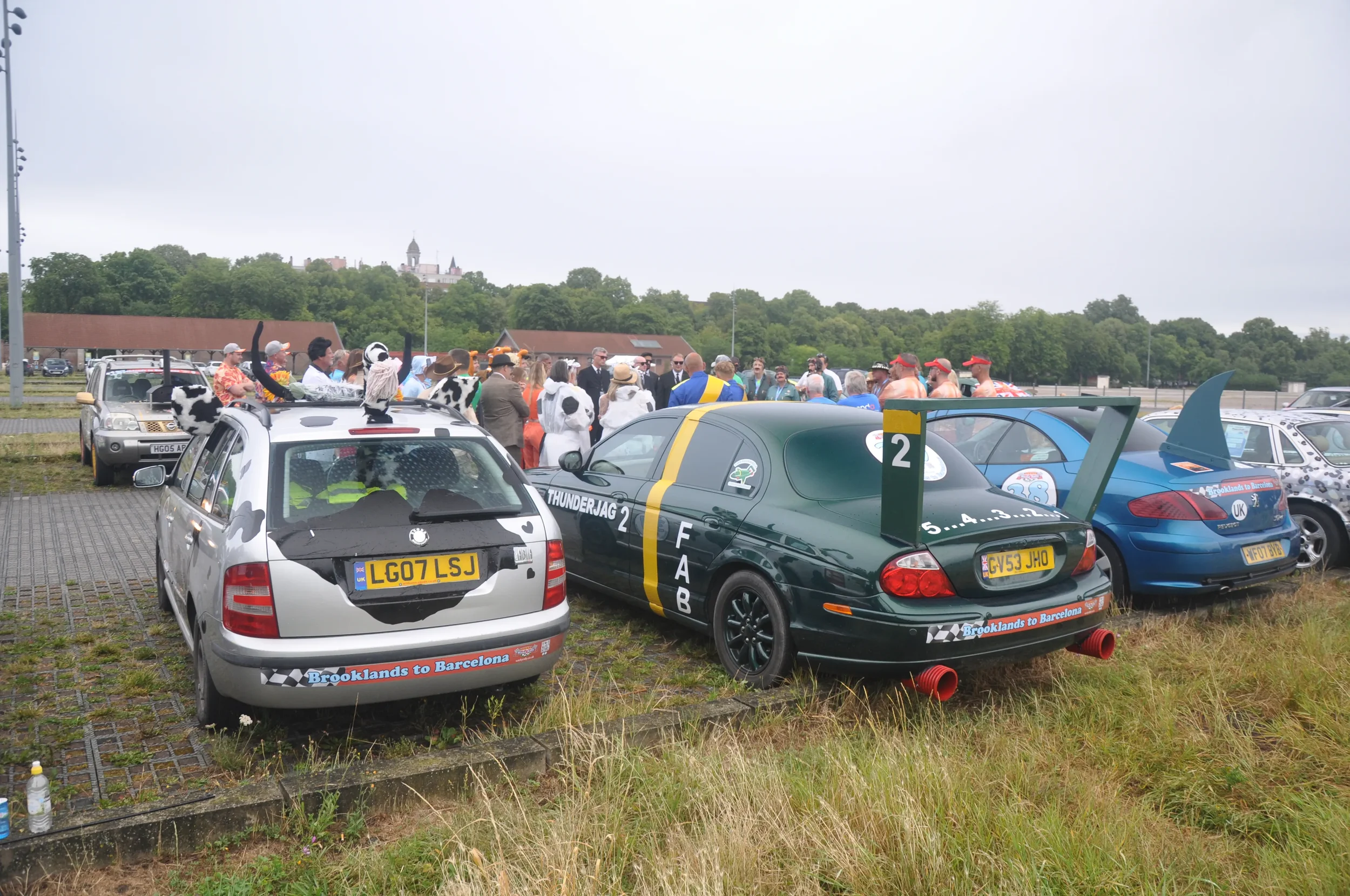 A group of people dressed in costumes and racing gear gathered around cars decorated with racing themes in an outdoor parking lot, with a hill and castle in the background.