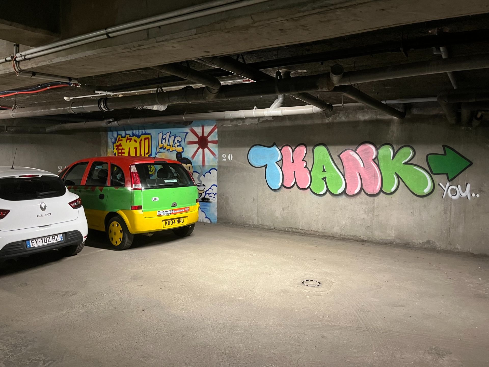Graffiti on a concrete wall with the words 'THANK YOU' in colorful letters, green and pink, with a green arrow pointing right. Two parked cars are nearby, one white and one painted with bright colors resembling a watermelon.