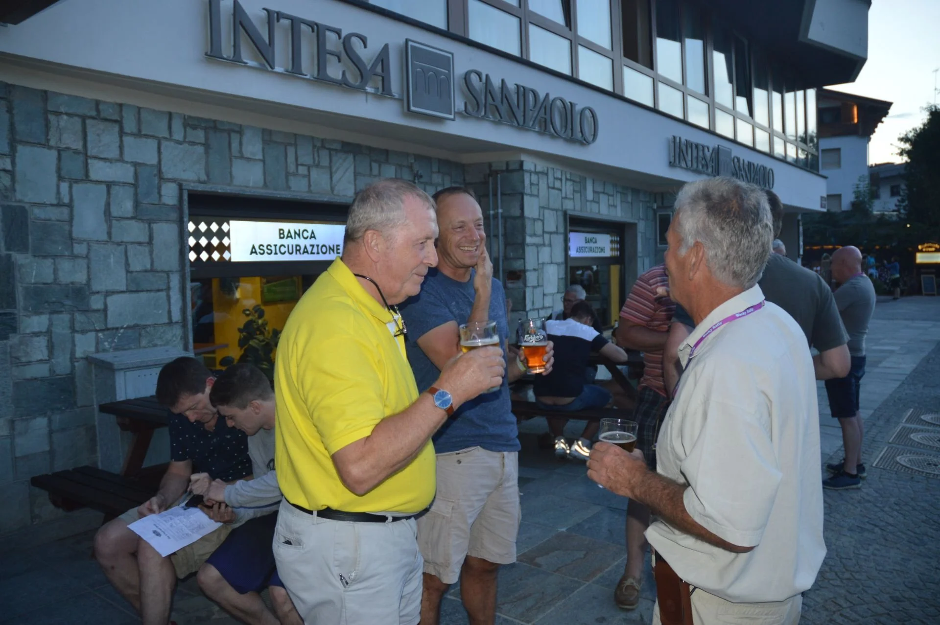Four men standing outside a building, holding beer glasses and engaging in conversation. Two young men are seated at a bench behind them, looking at their phones. The building has a sign reading 'BANCA ASSICURAZIONE' with a stone exterior and large w