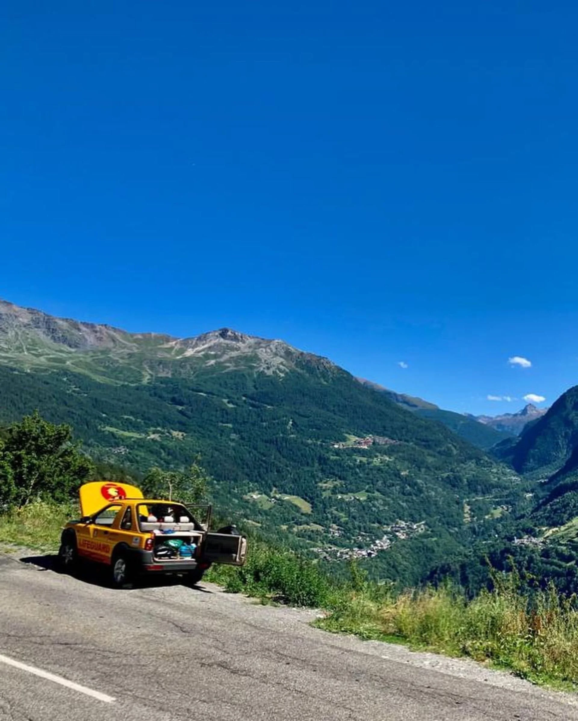 A yellow lifeguard car parked on the side of a mountain road with an open trunk, overlooking lush green mountains and a valley under a clear blue sky.